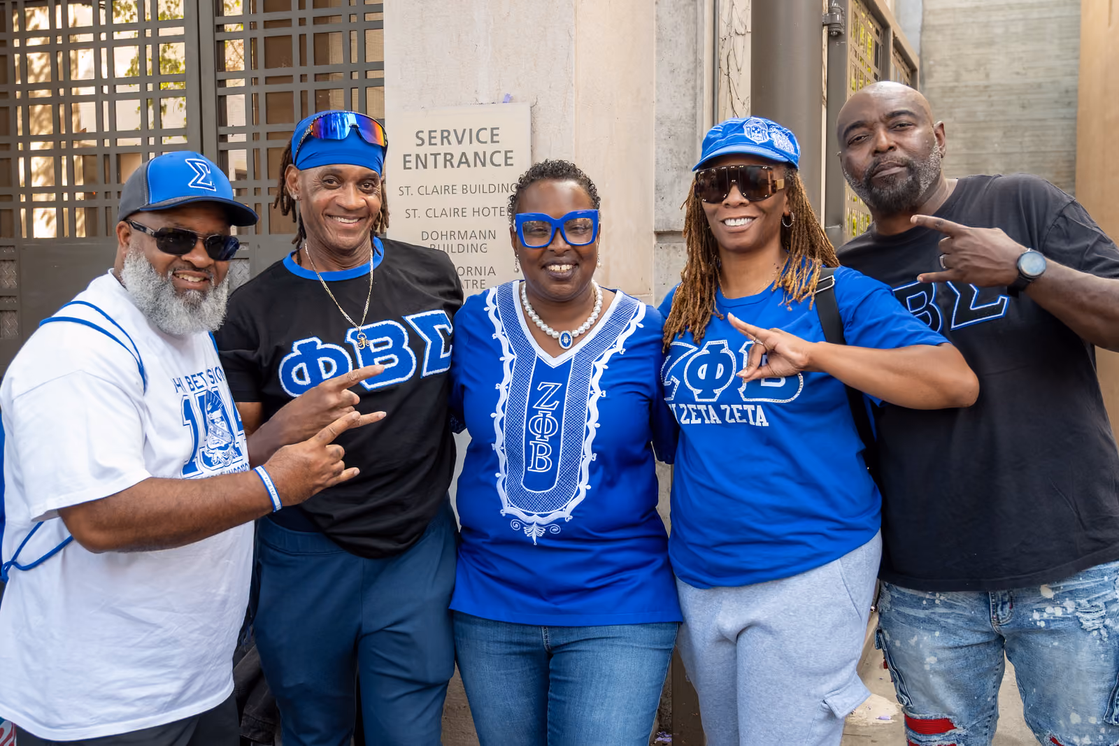 Five festival guests wearing blue, black and white Greek-letter clothing pose together beside a building