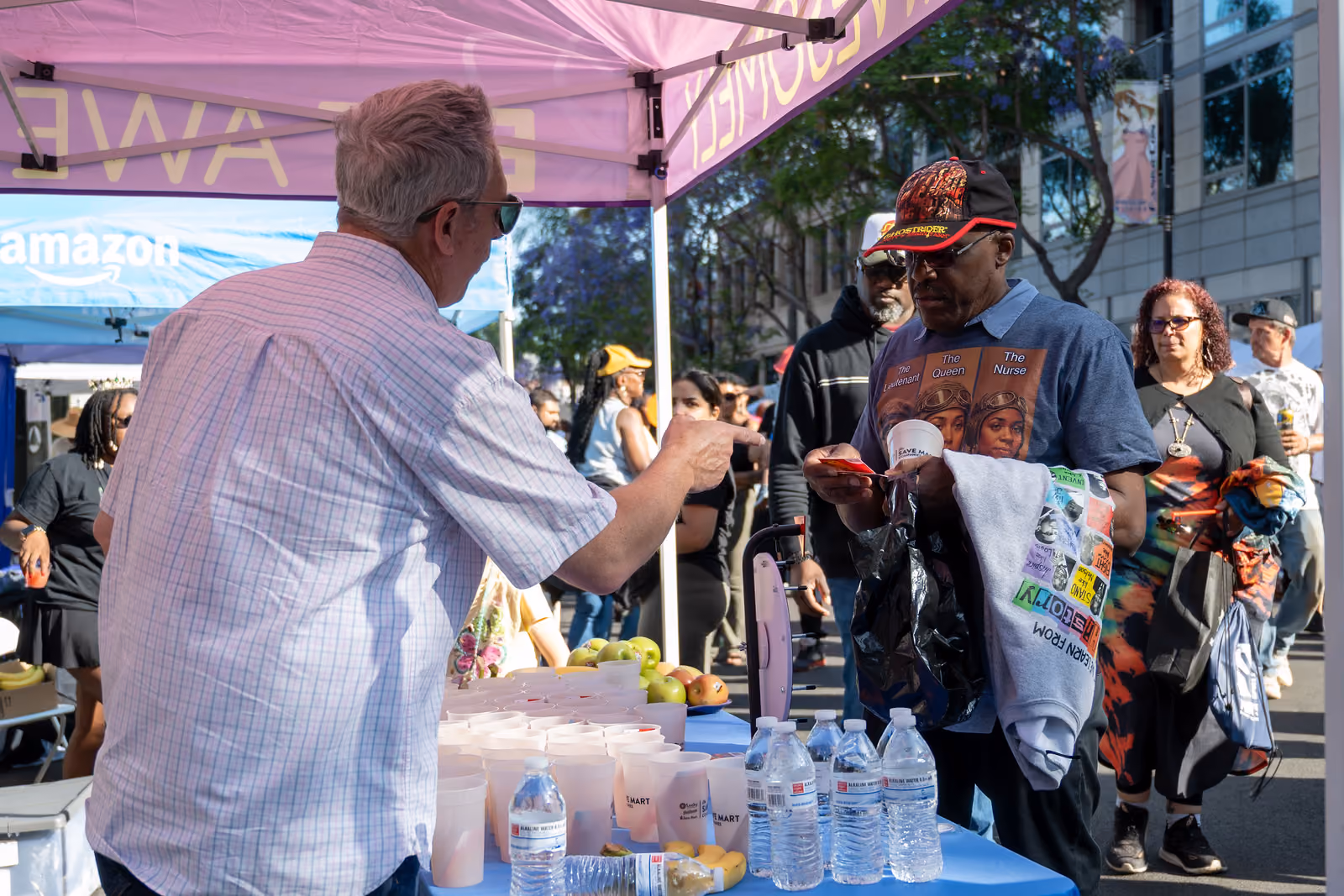 A booth attendant gestures toward a visitor across a table stocked with water bottles, cups and fruit