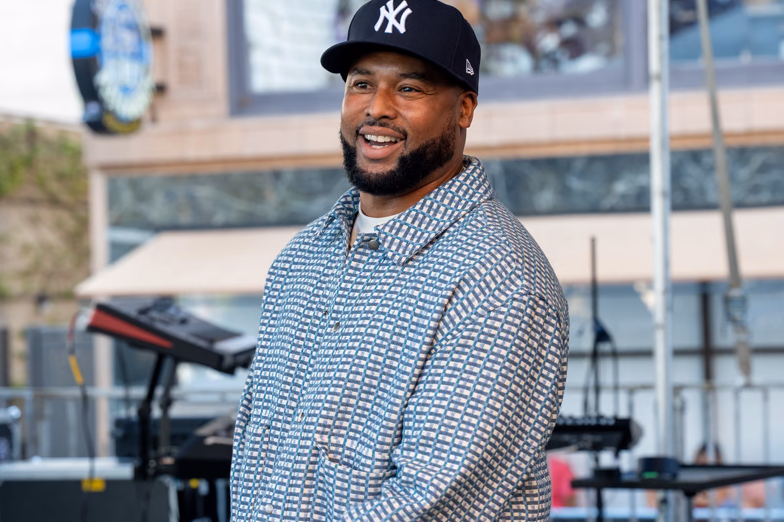 A smiling man in a Yankees cap and patterned jacket stands on an outdoor stage near musical equipment