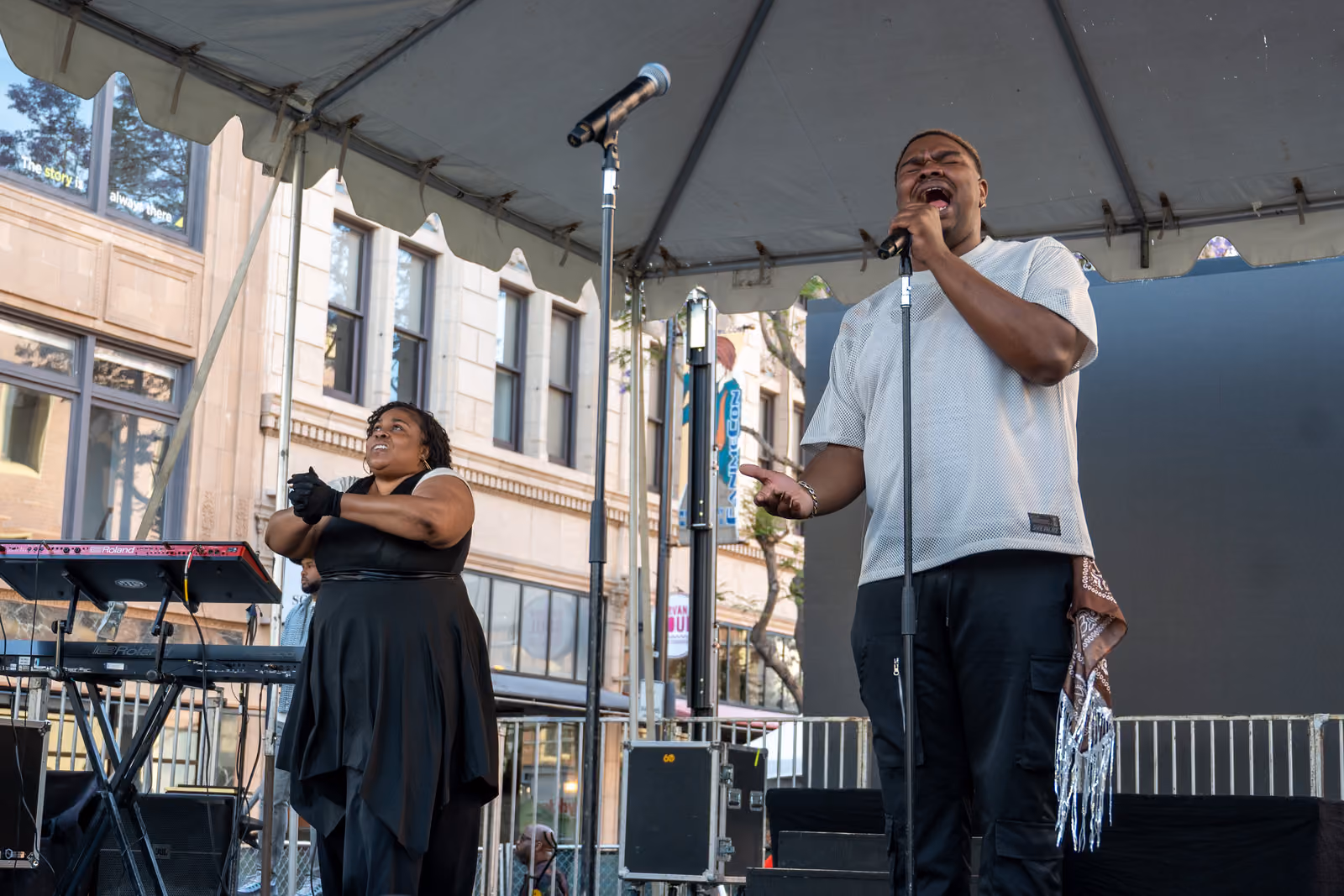 A singer in a white shirt performs into a microphone beside a woman gesturing near keyboards onstage