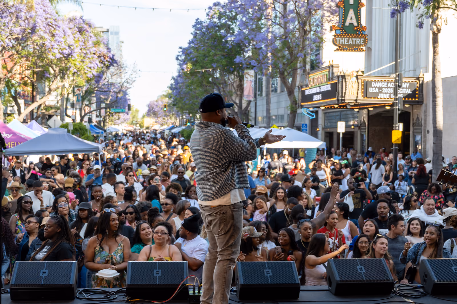 A performer holds a microphone and gestures toward a large street crowd from the outdoor stage