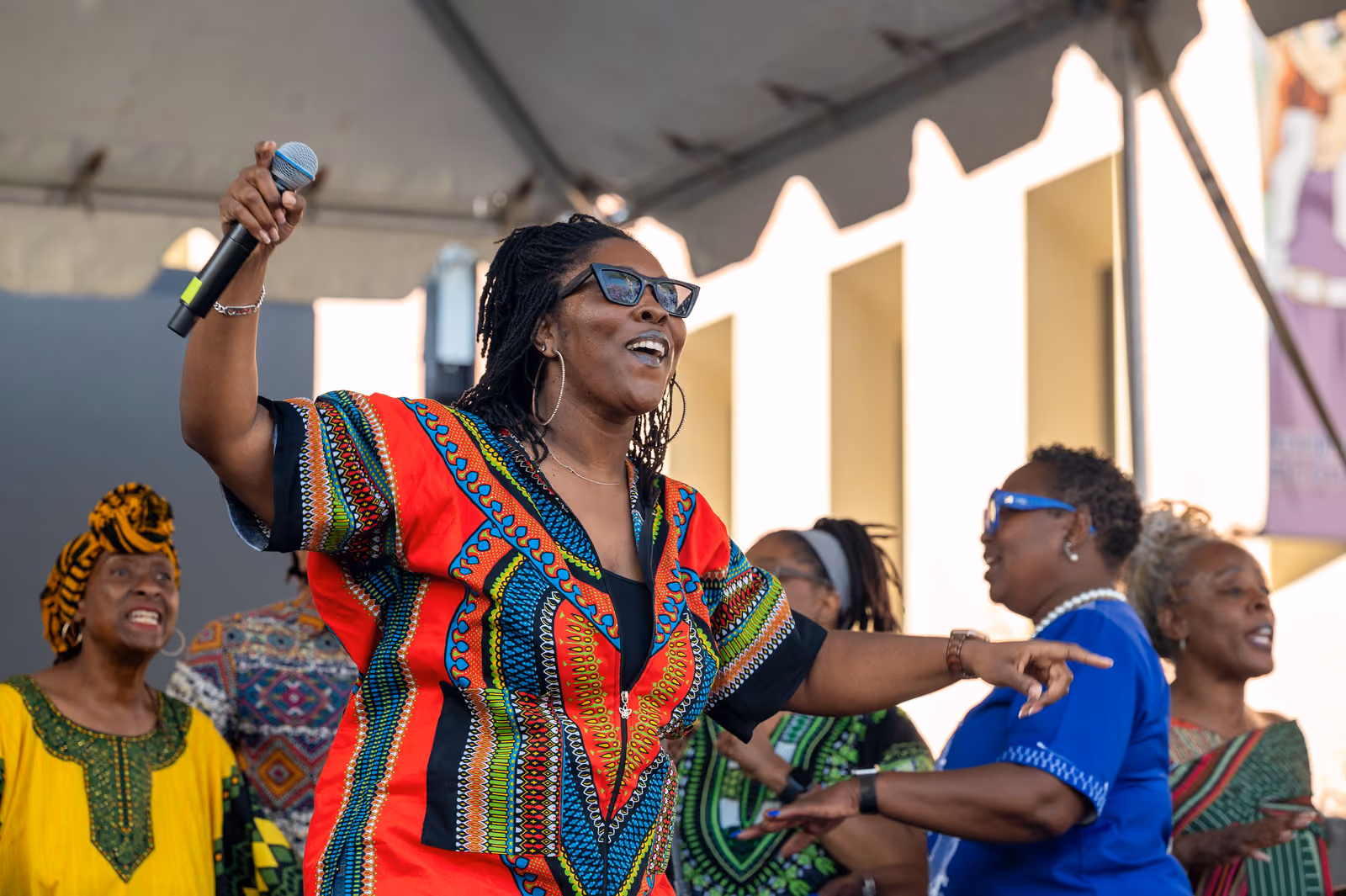 A smiling singer in a red patterned shirt raises a microphone while fellow singers stand behind her