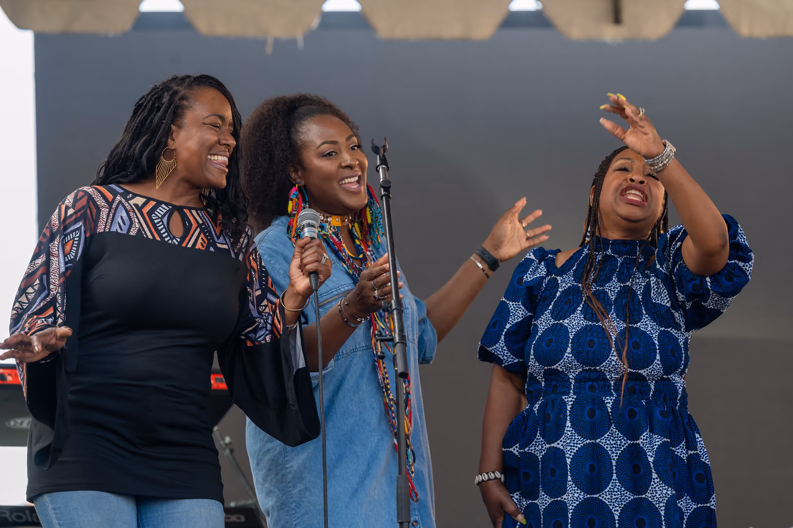 Three singers perform together on a covered stage, with one holding a microphone and another raising a hand