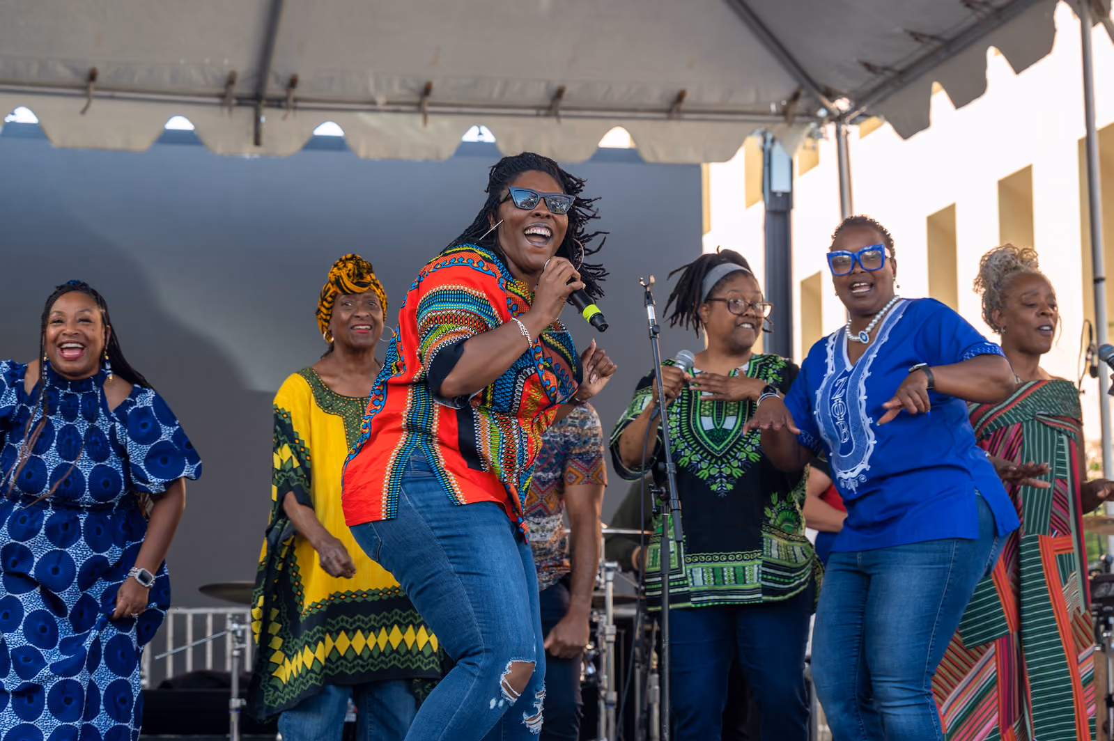 Singers in colorful patterned clothing smile and move together beneath the outdoor stage canopy