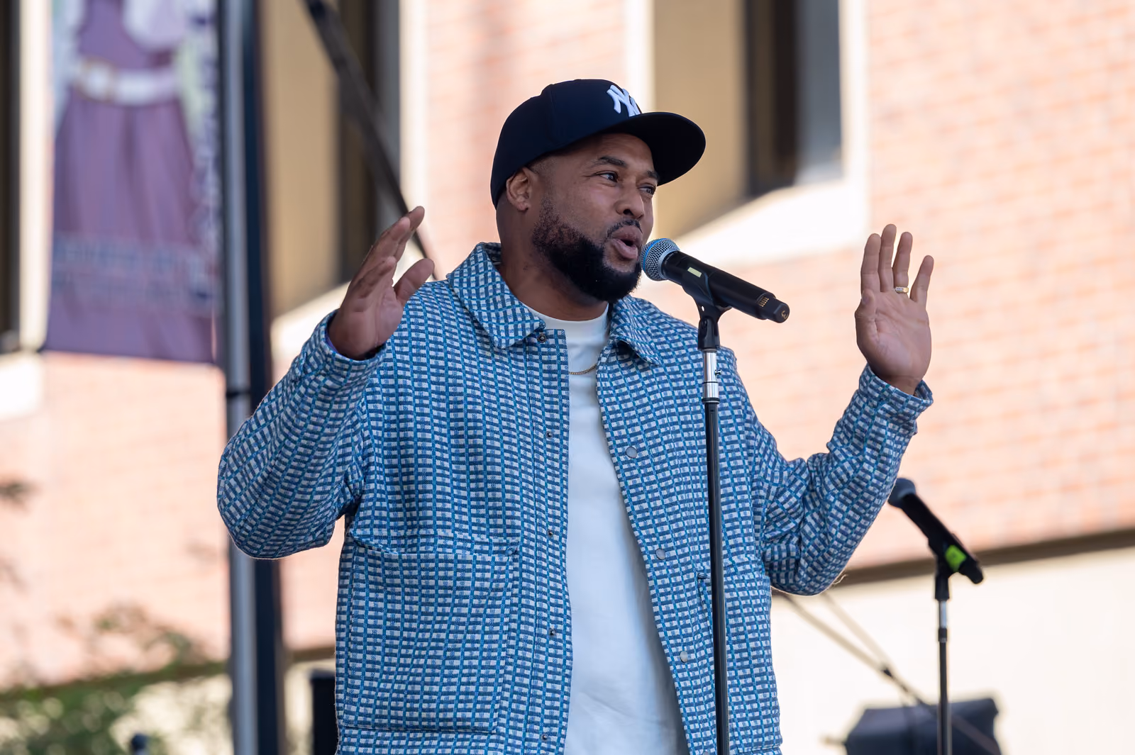 A speaker in a Yankees cap raises both hands while addressing a microphone on the outdoor stage