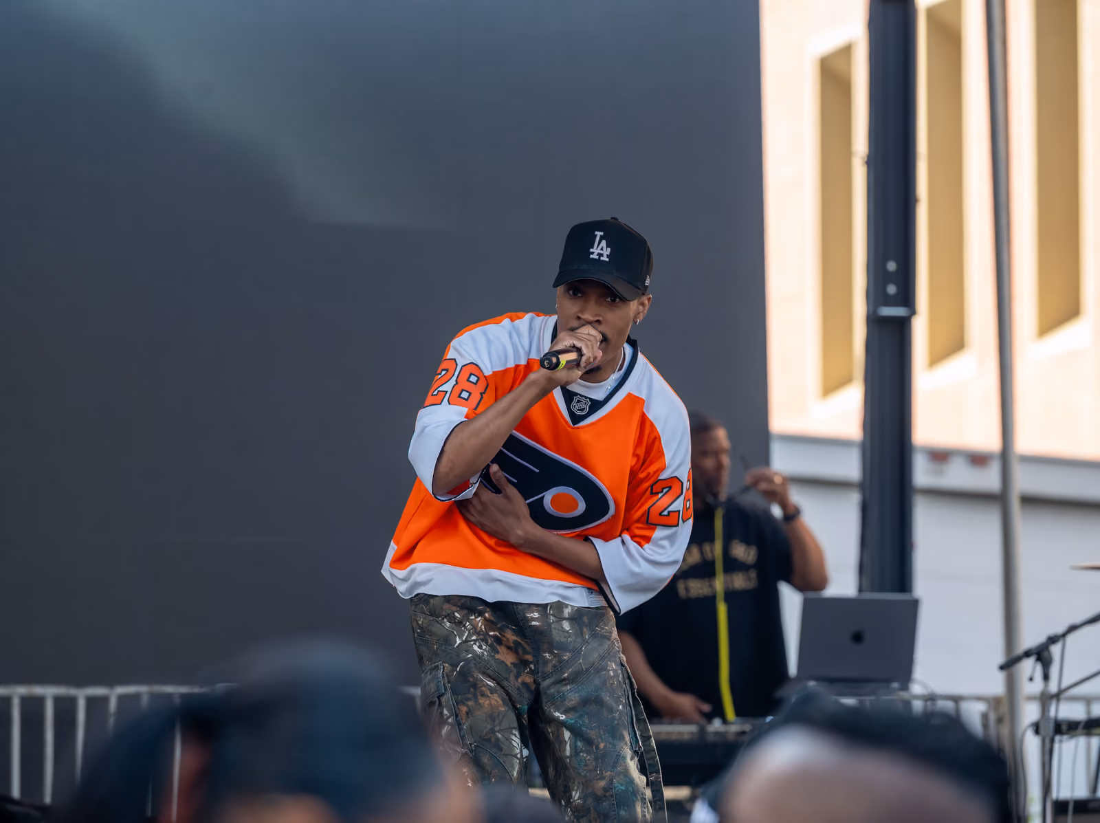 A rapper in an orange hockey jersey and black cap performs with a handheld microphone onstage
