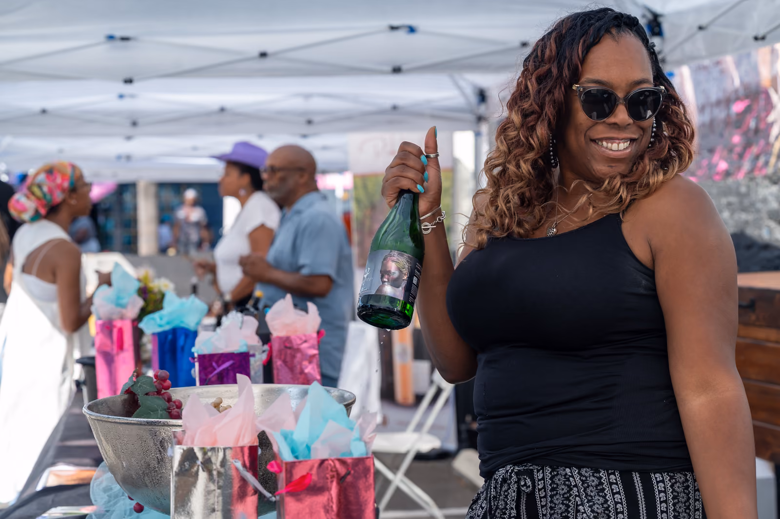 A smiling guest in sunglasses holds up a green bottle beside colorful gift bags under a canopy