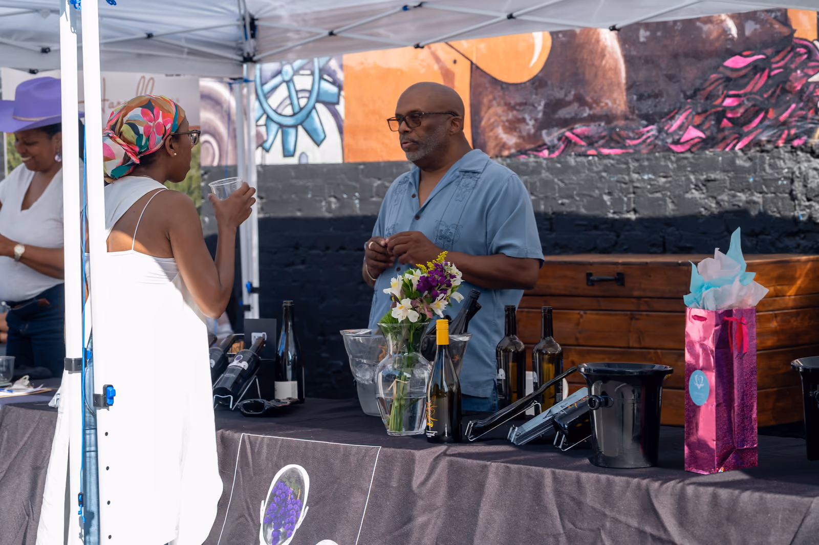 A visitor holding a tasting cup talks with a vendor behind a table displaying wine bottles and flowers