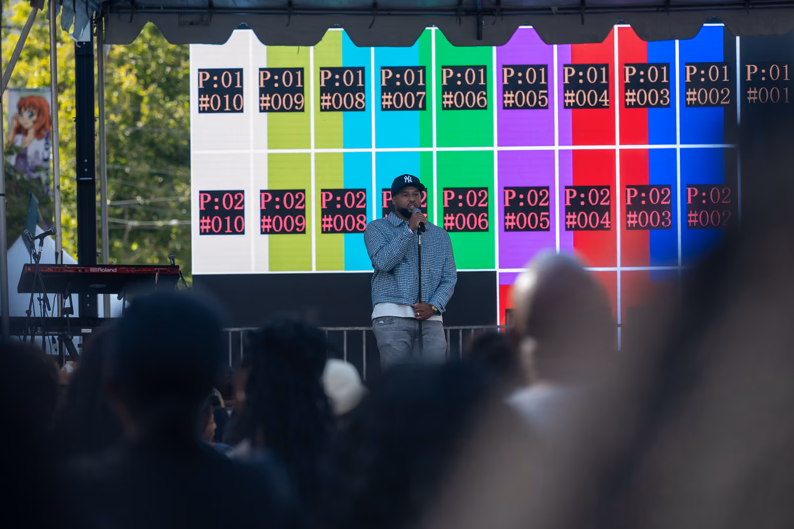 A performer in a baseball cap holds a microphone before a colorful screen, with the crowd in front