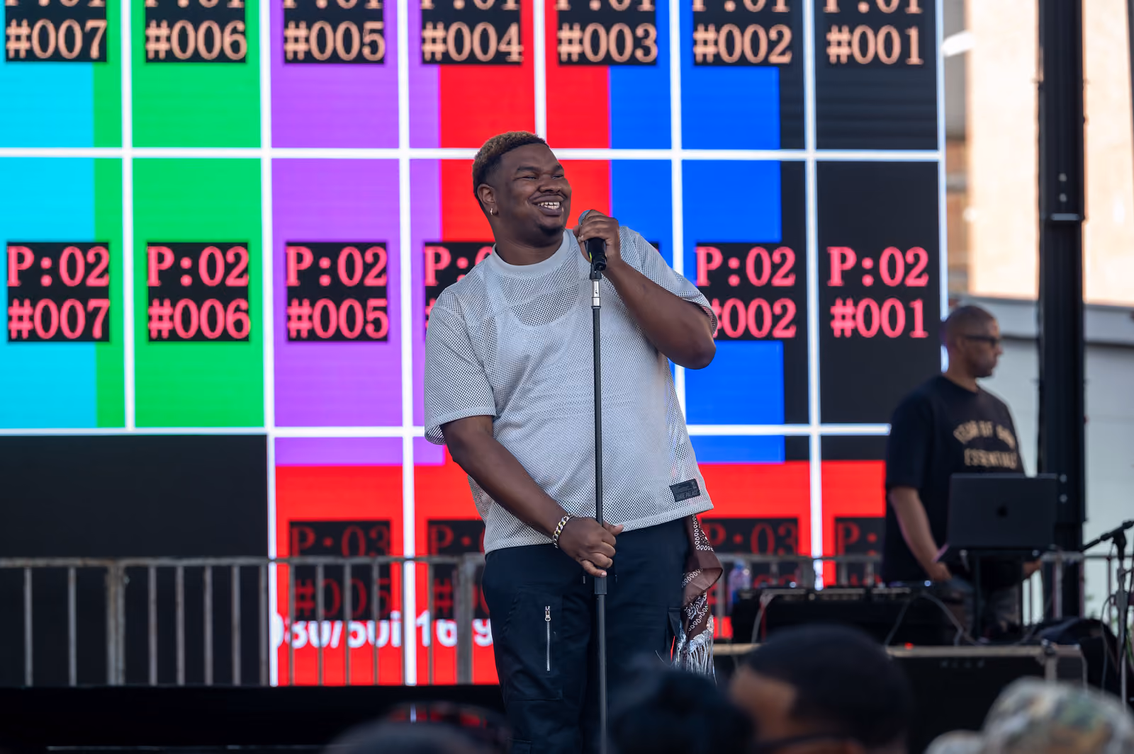A smiling performer holds a microphone stand onstage in front of a bright multicolored screen