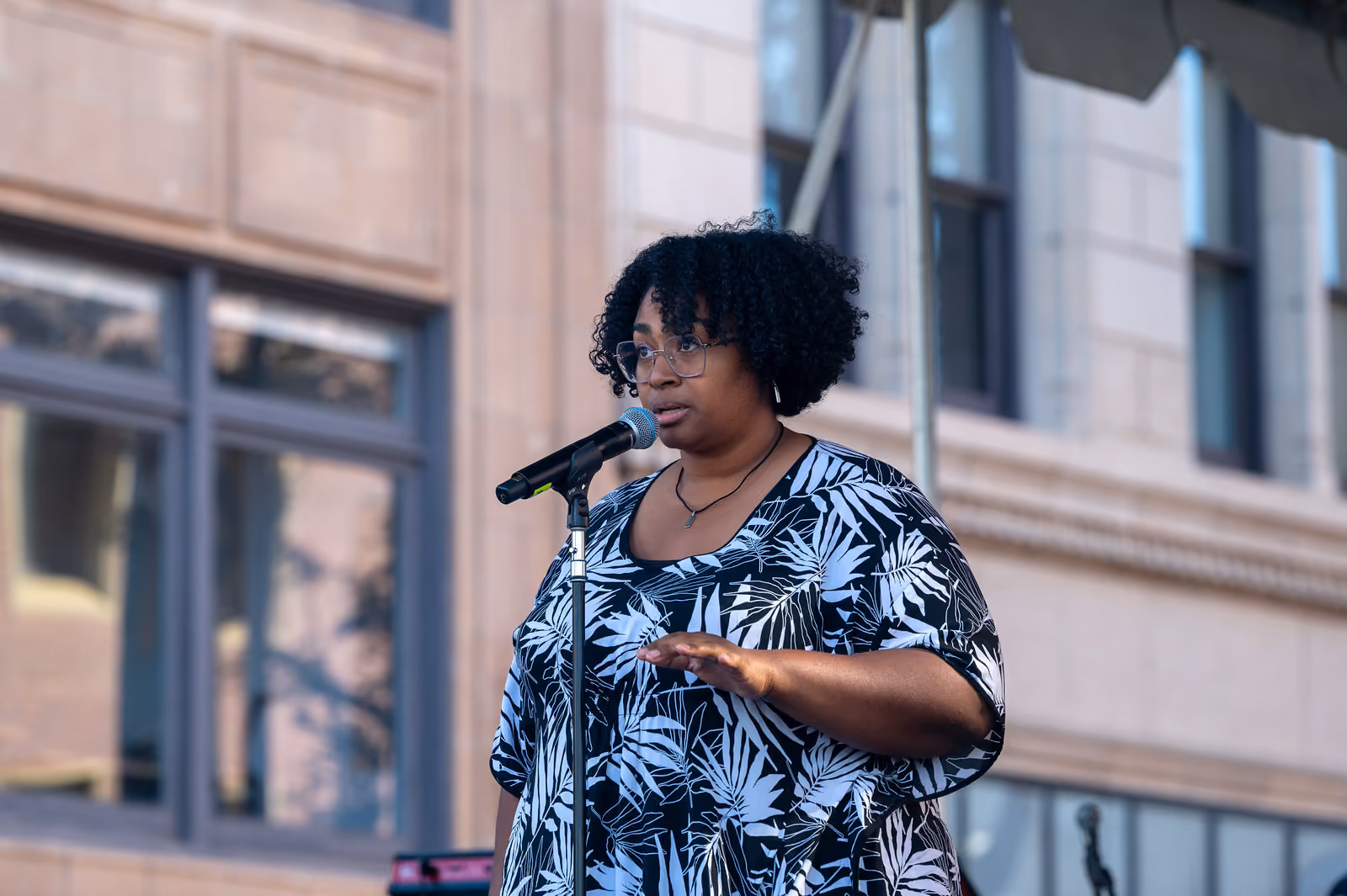 A speaker in a black and white leaf-patterned top gestures at a microphone on an outdoor stage