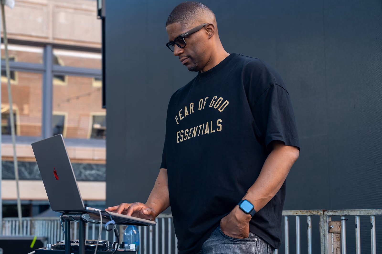 A man in sunglasses and a black T-shirt operates a laptop on a stand beside the outdoor stage