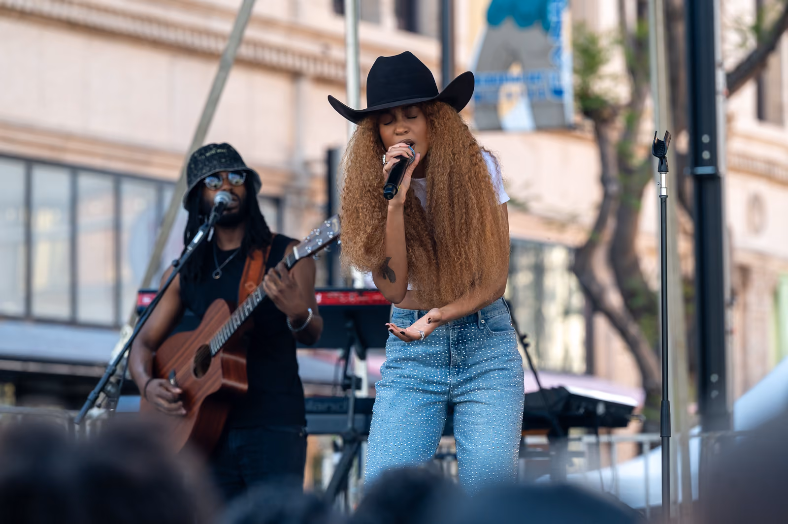 A singer in a black cowboy hat leans into a handheld microphone while a guitarist plays beside her