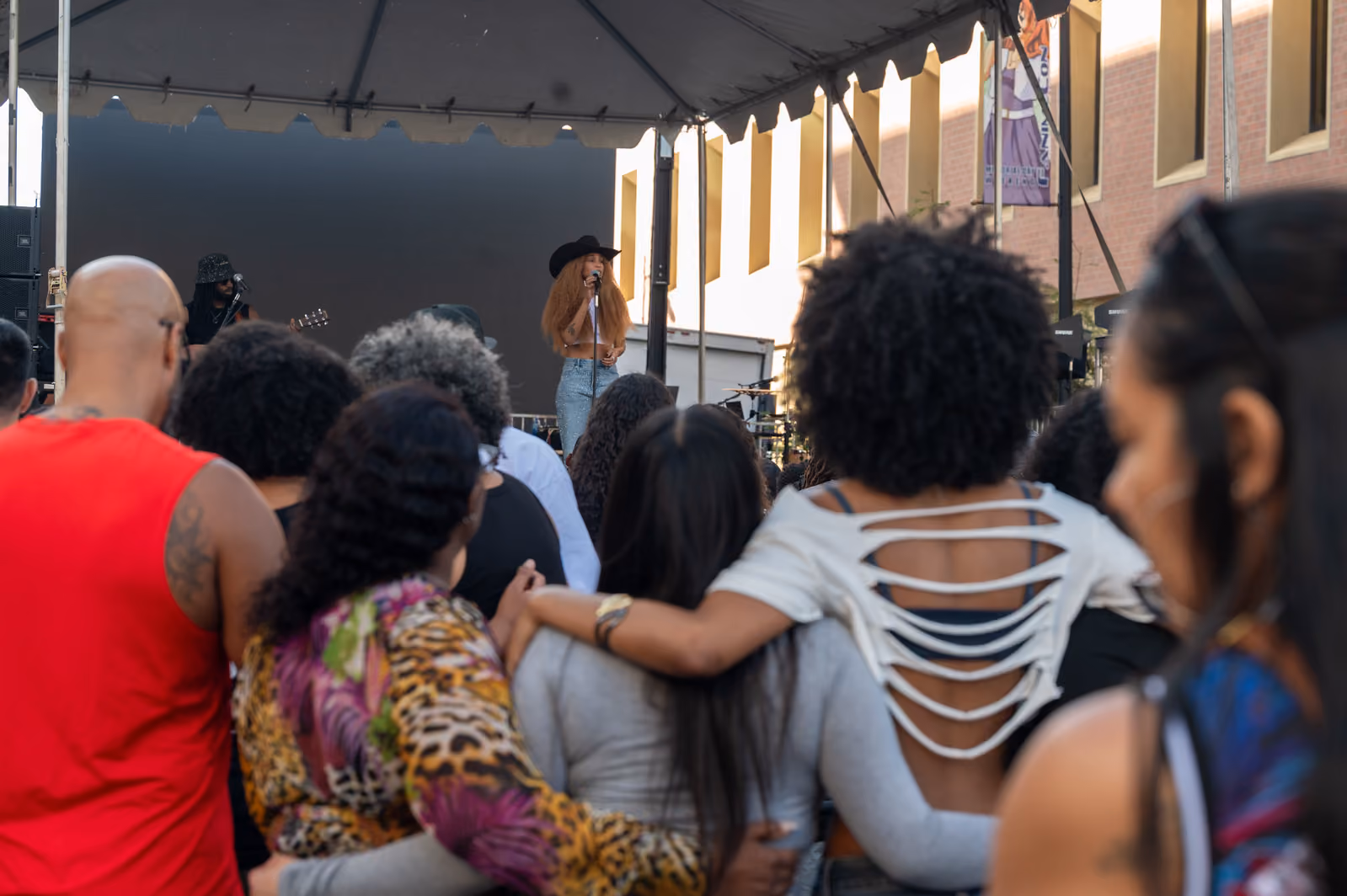 Festivalgoers stand with arms around one another as they watch a singer perform beneath a stage canopy