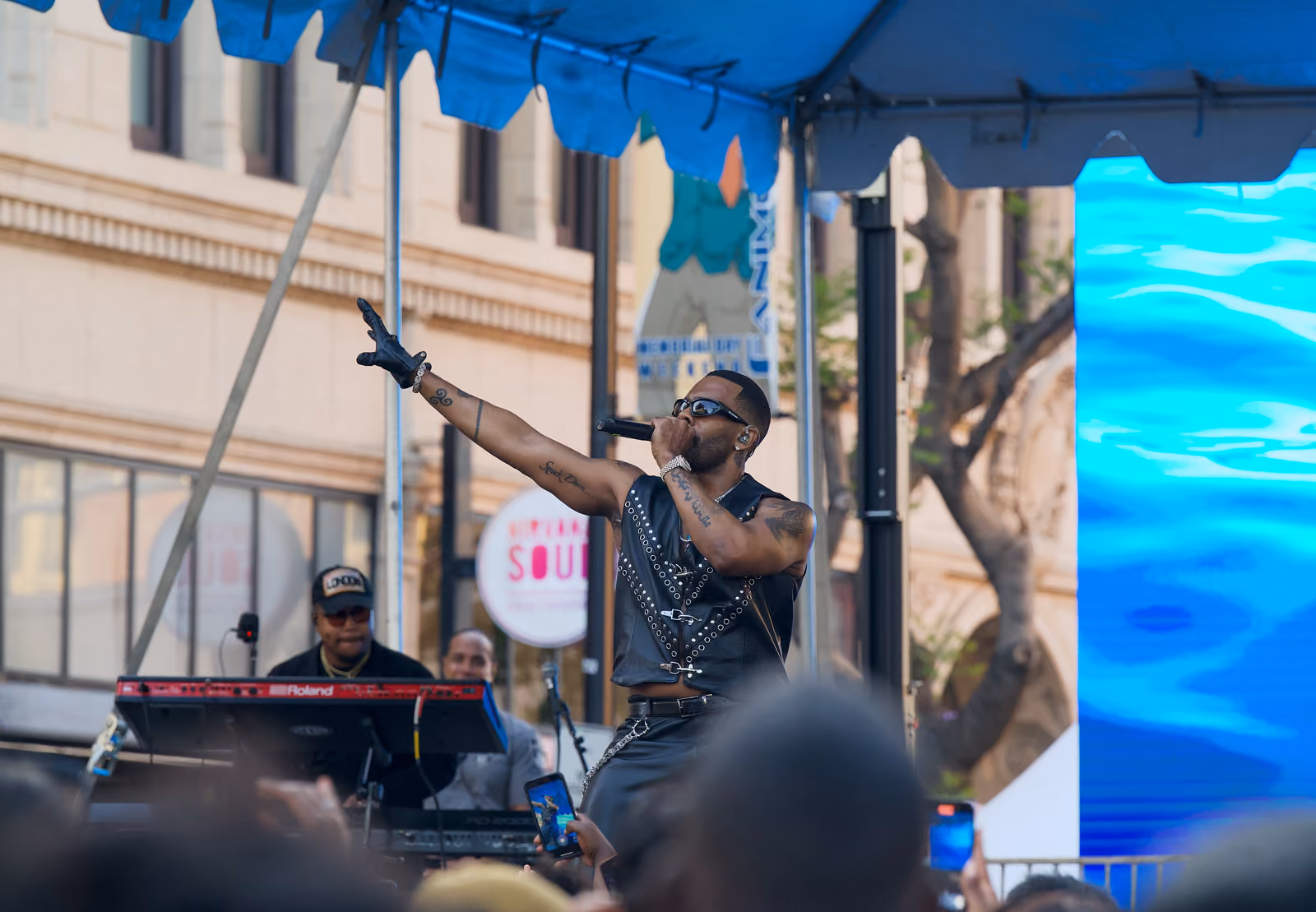 A singer in a black leather vest reaches one arm toward the crowd while a keyboardist plays behind him
