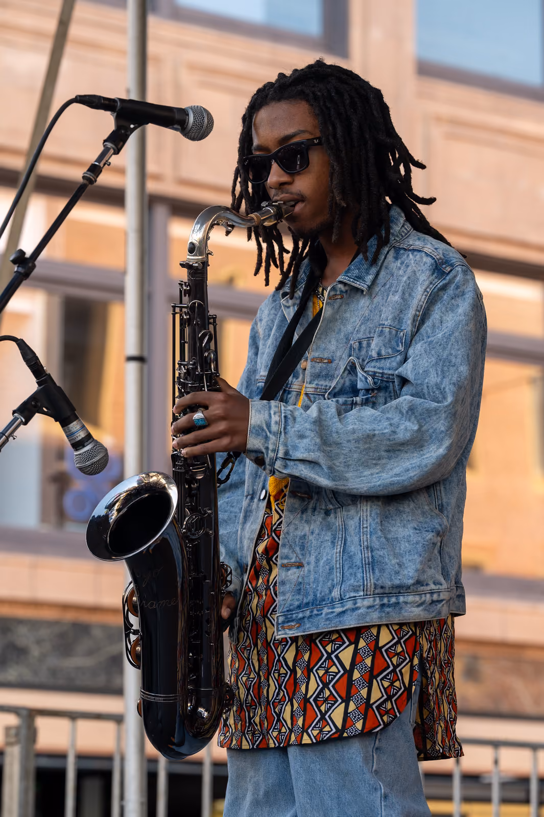A musician in sunglasses and a denim jacket plays a black saxophone beside microphones onstage