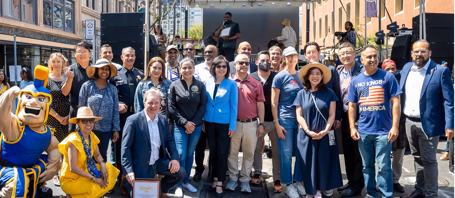 A large group poses in front of the festival stage beside a blue and yellow mascot and a framed certificate