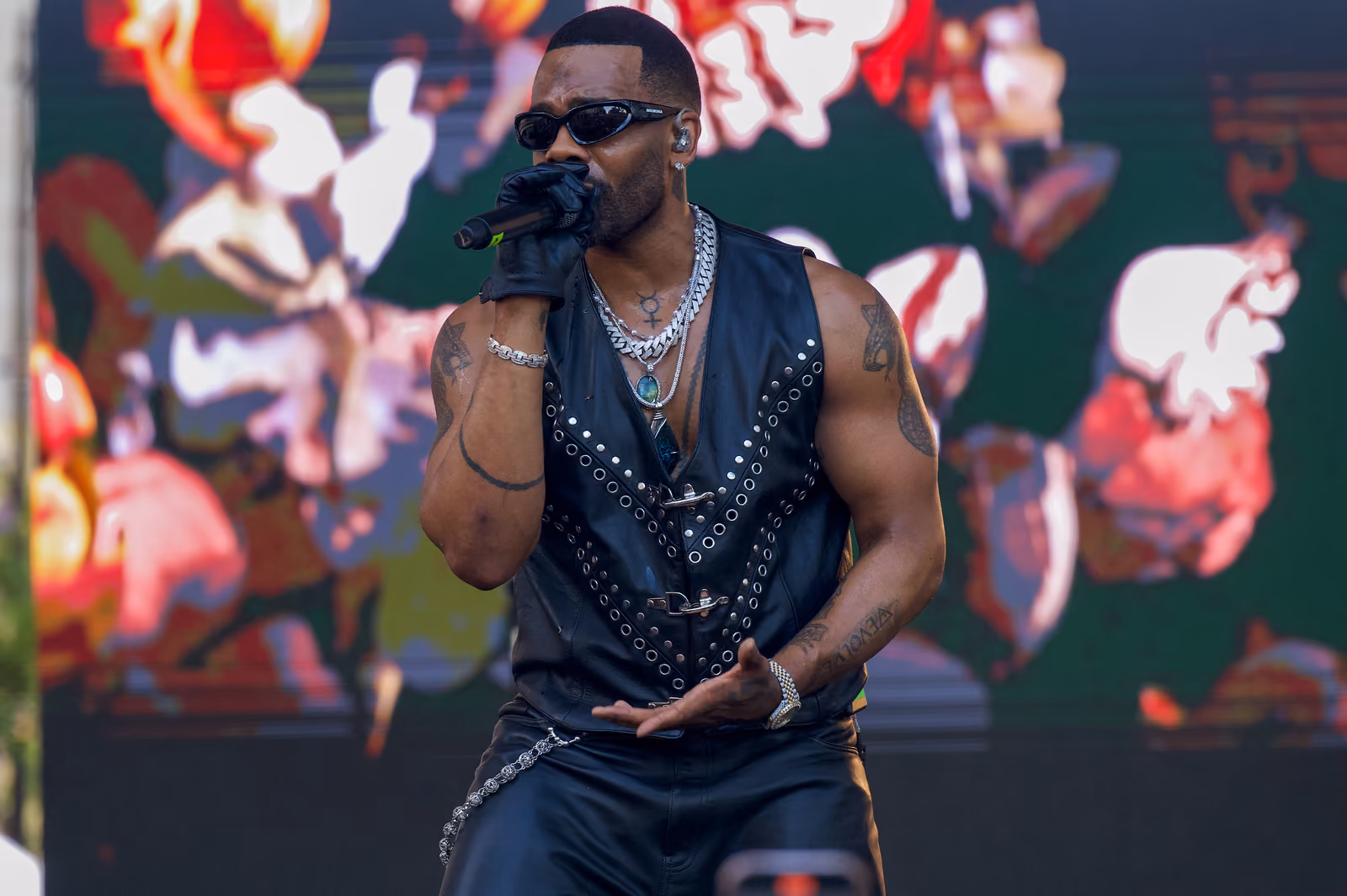 A singer in a studded leather vest sings into a microphone before a screen filled with colorful visuals