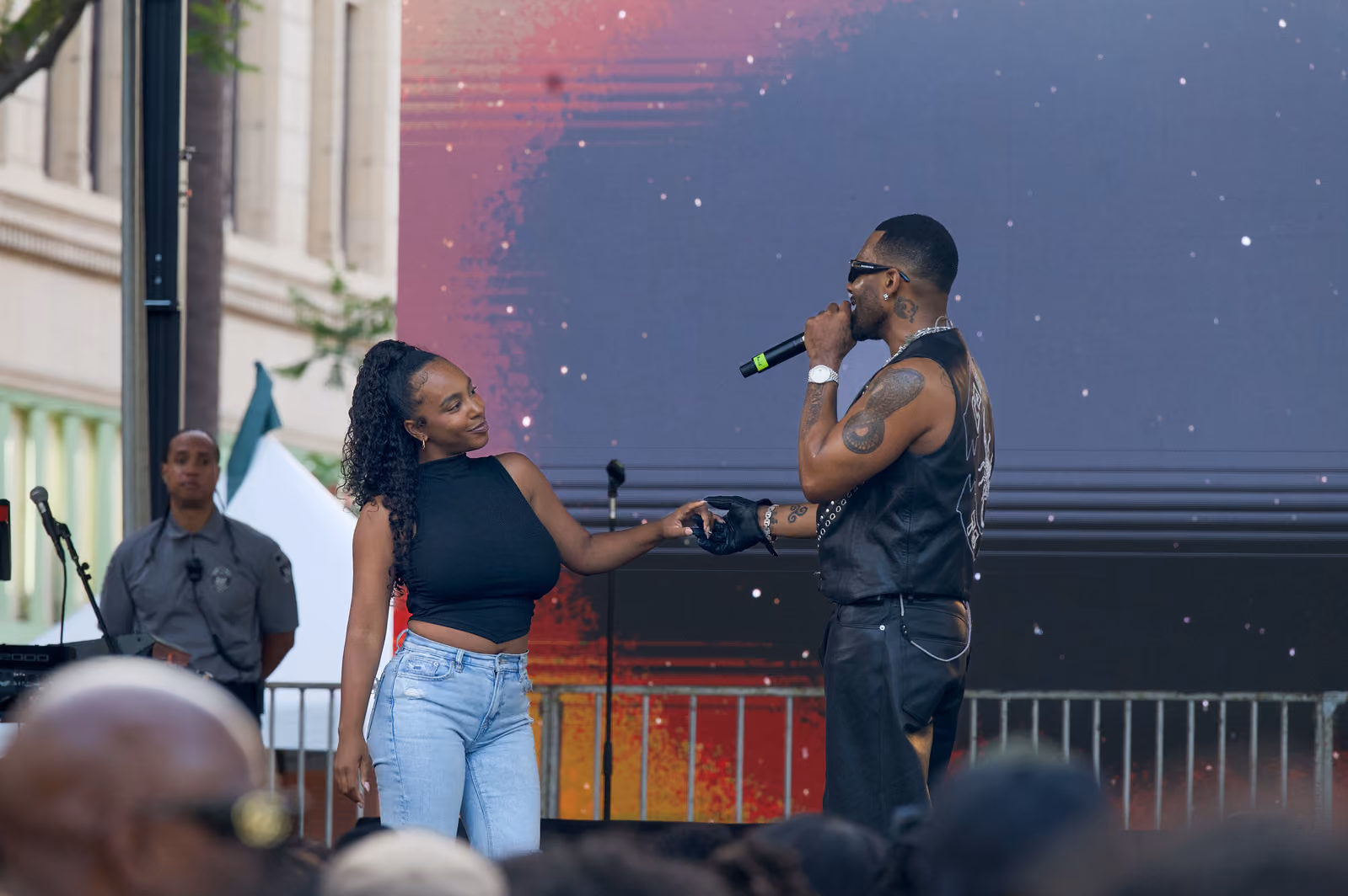 A singer holds hands with a guest in a black sleeveless top and light jeans on the outdoor stage