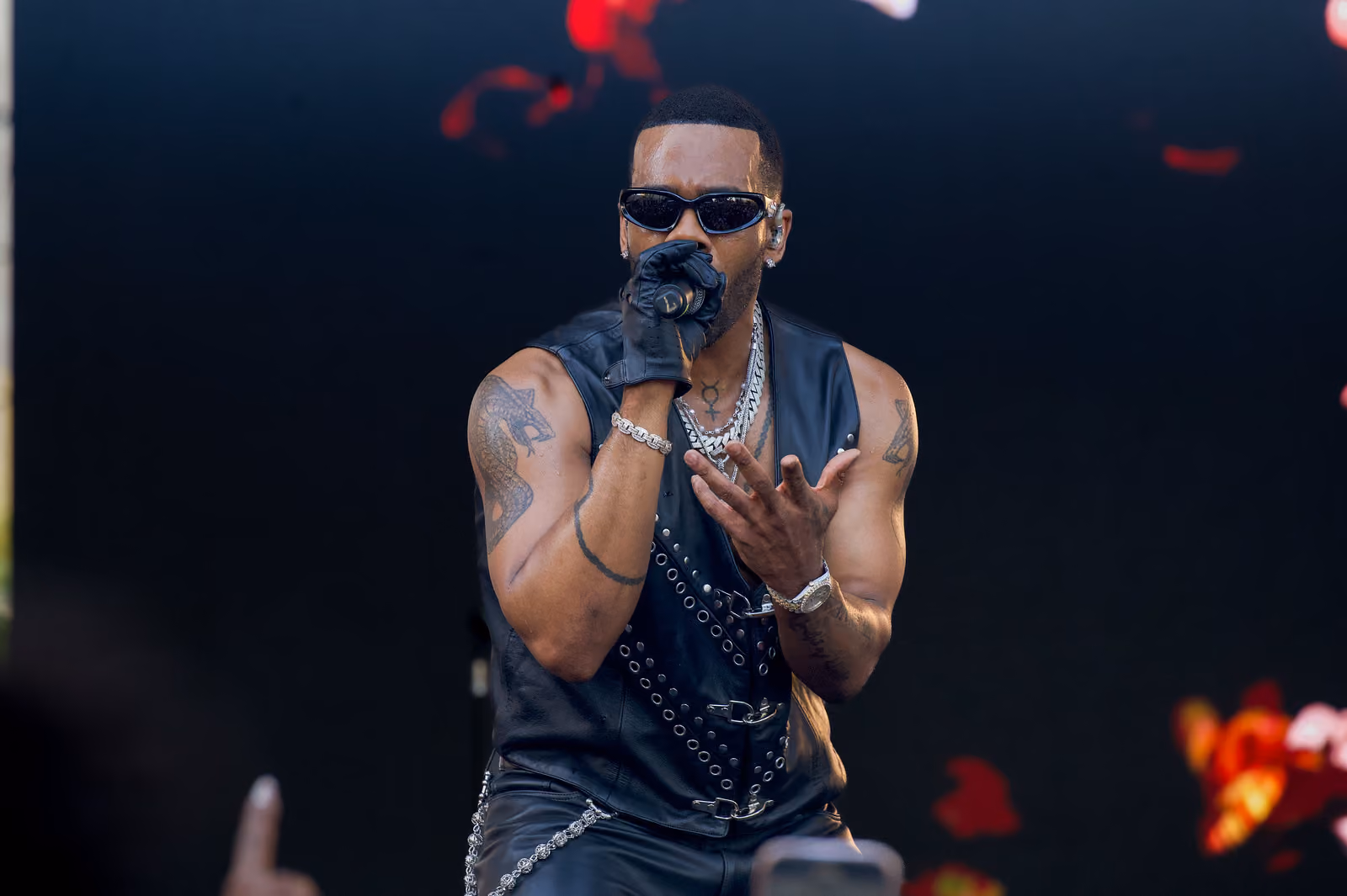 A singer in sunglasses holds a microphone in a gloved hand and extends his other palm toward the crowd