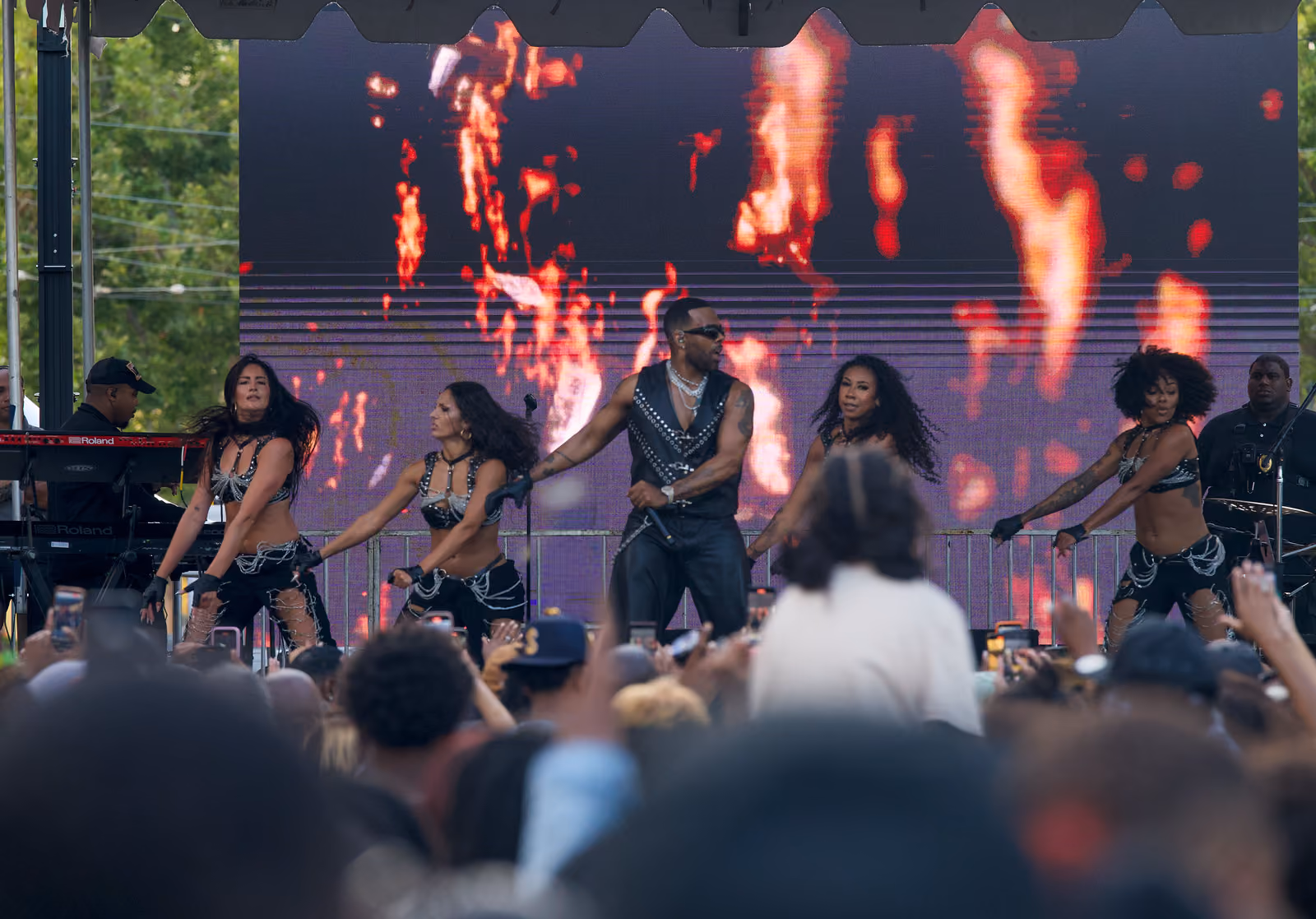 A singer and four dancers perform synchronized moves before a flame-filled screen as the crowd watches