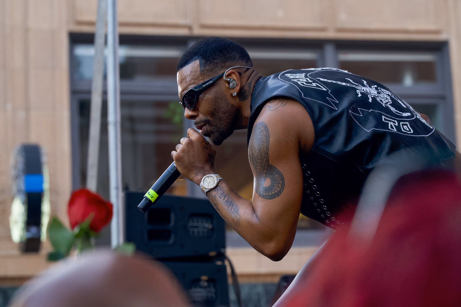 A singer in sunglasses bends toward the audience with a microphone as a red rose rises in the foreground
