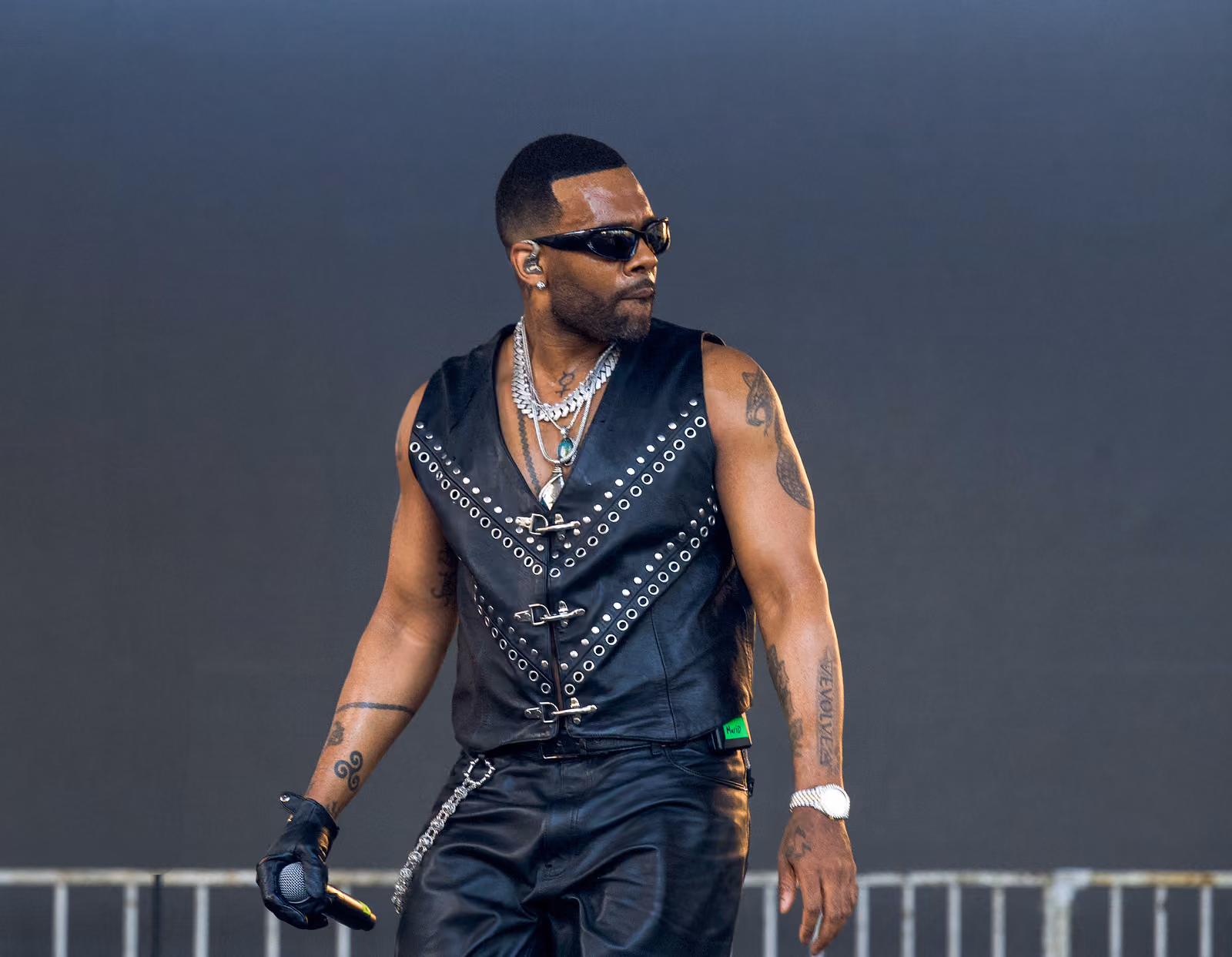 A performer in sunglasses and a studded black leather vest looks to the side with a microphone lowered