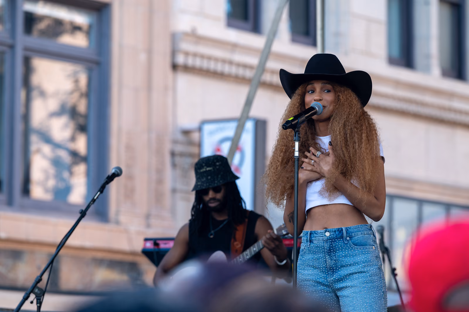 A singer in a black cowboy hat holds a hand to her chest at a microphone, with a guitarist behind her