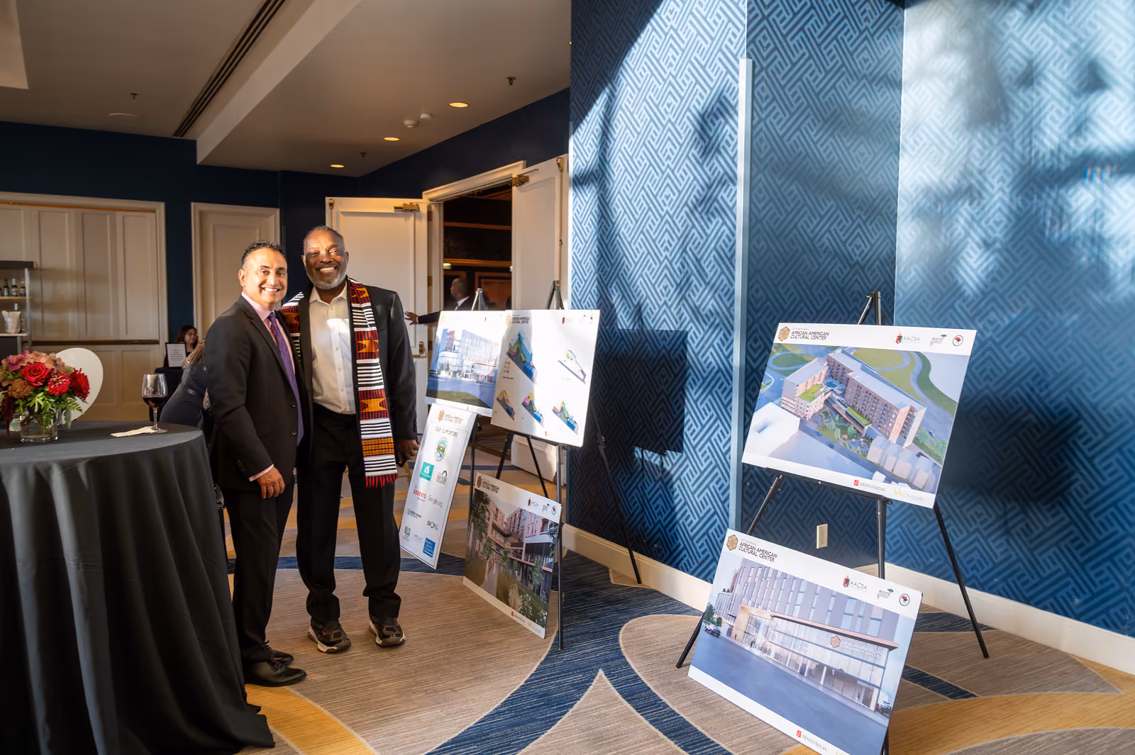 Two guests in suits pose beside architectural renderings displayed on easels in the gala foyer