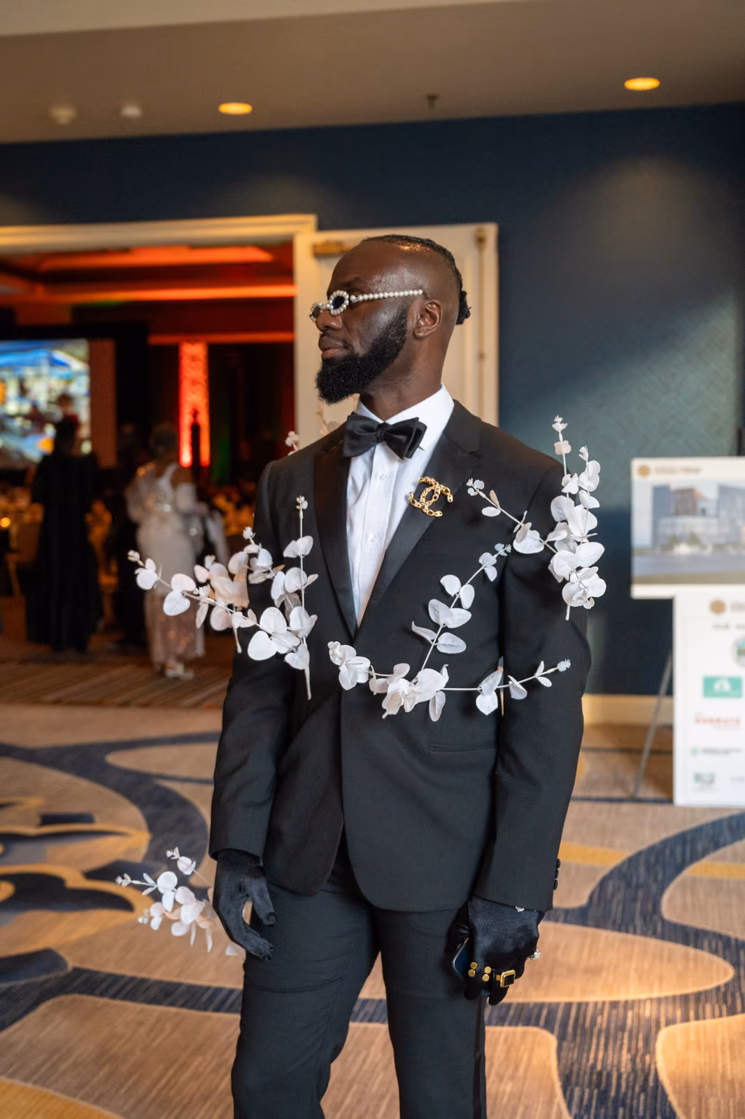 A guest in a black tuxedo with white leafy decorations and jeweled glasses poses in the foyer