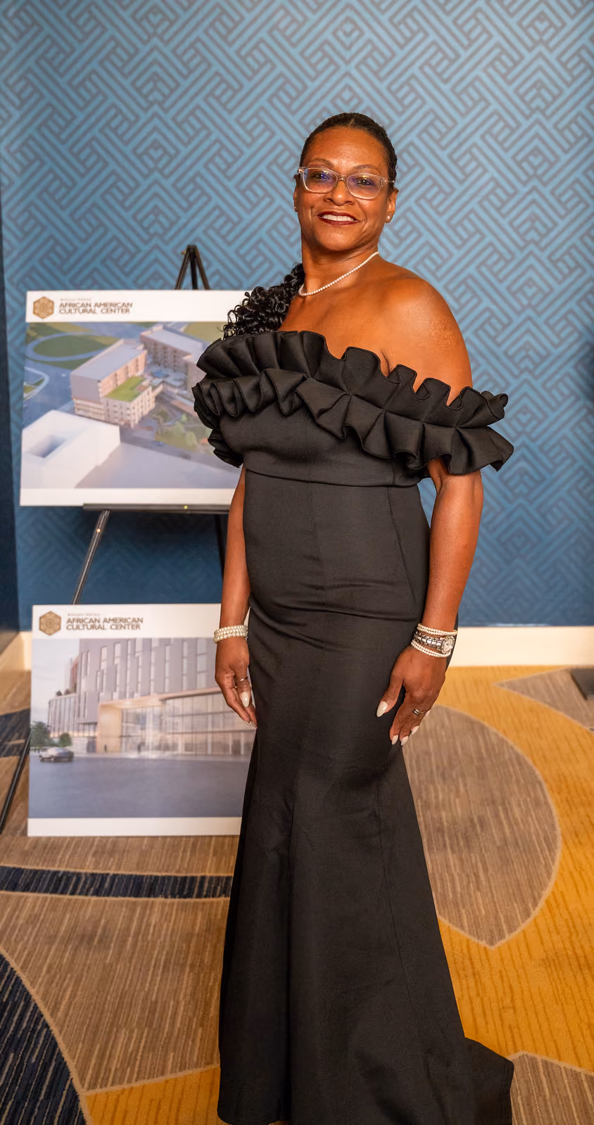 A guest in a black ruffled gown poses beside architectural renderings on easels in the gala foyer