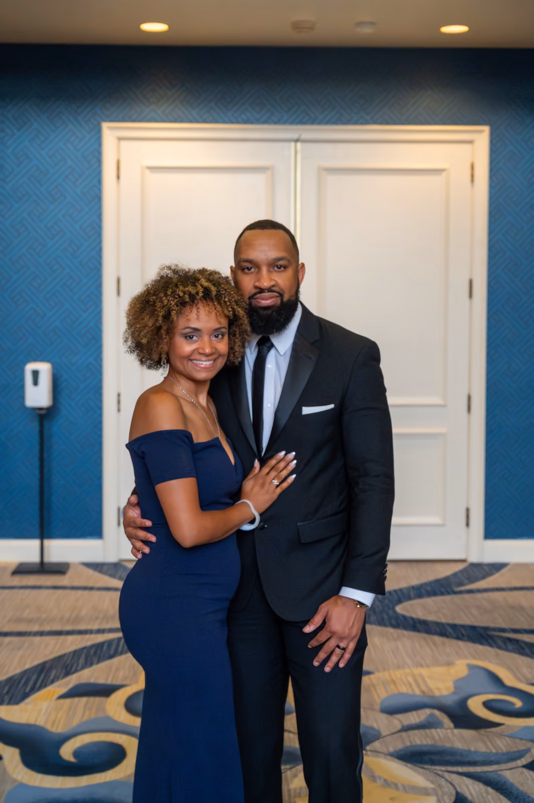 Two guests in a navy gown and black suit pose with their arms around each other in front of white doors