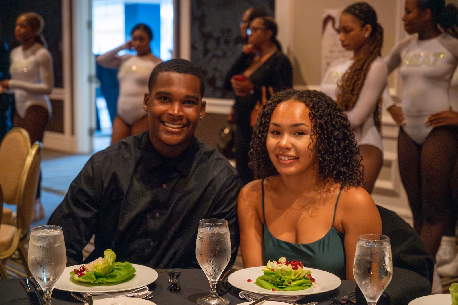 Two guests sit at a table set with salads and water glasses as dancers stand behind them