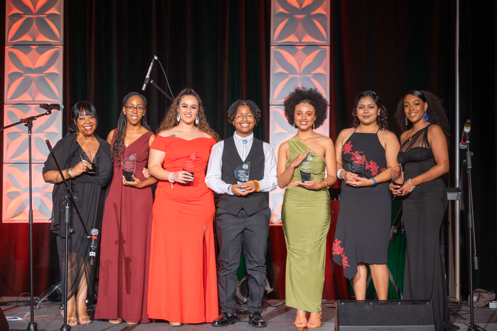 Seven guests stand together onstage, most holding clear awards, between illuminated decorative panels