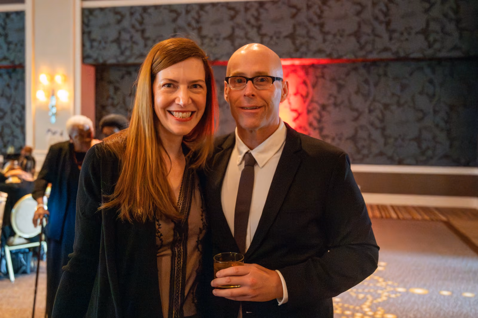 Two smiling guests in formal attire pose together in the ballroom, one holding a drink