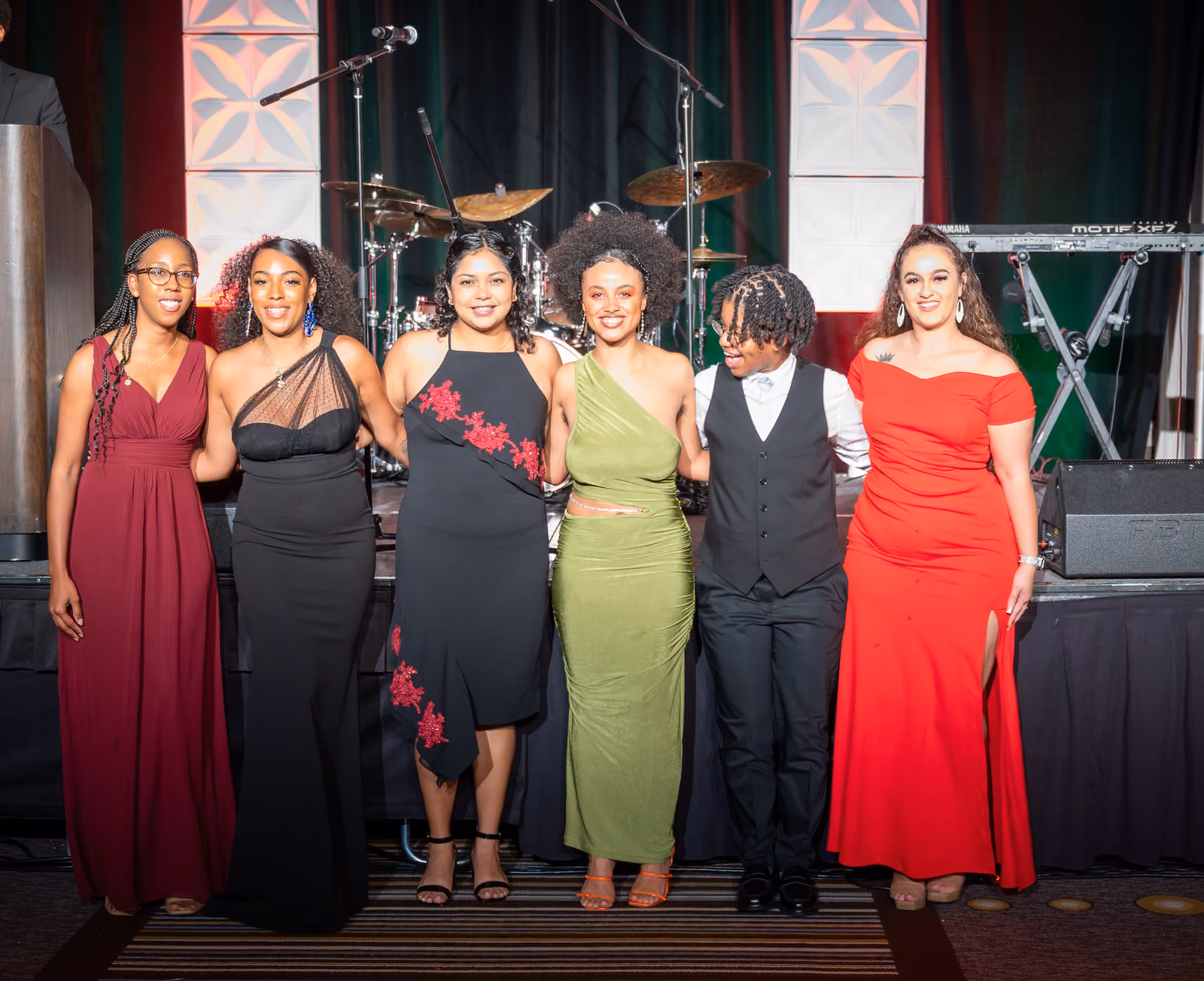 Six guests in formal attire pose in front of the stage with a drum kit and keyboard behind them