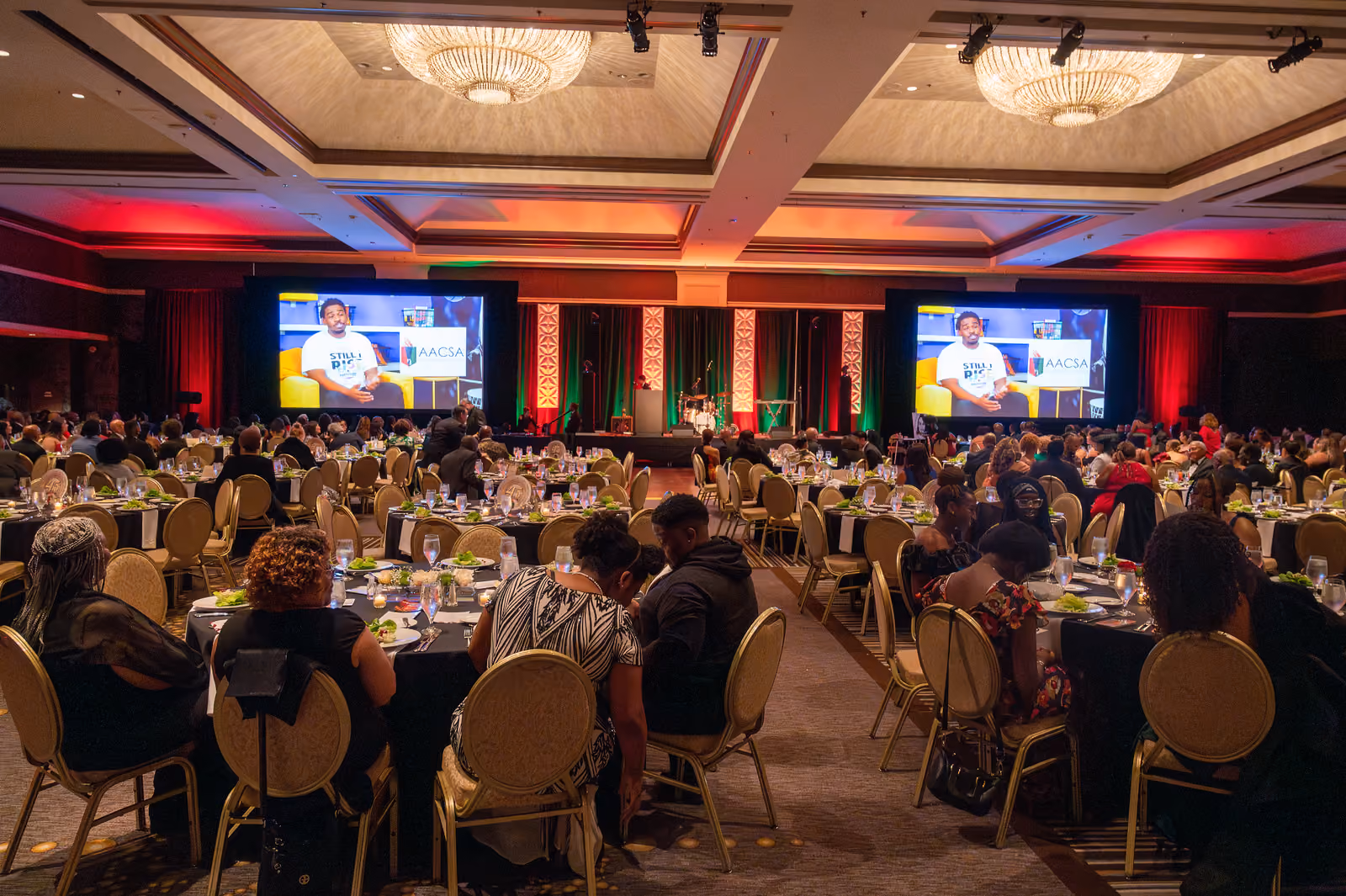 Guests fill round banquet tables facing a stage flanked by two large video screens in the ballroom