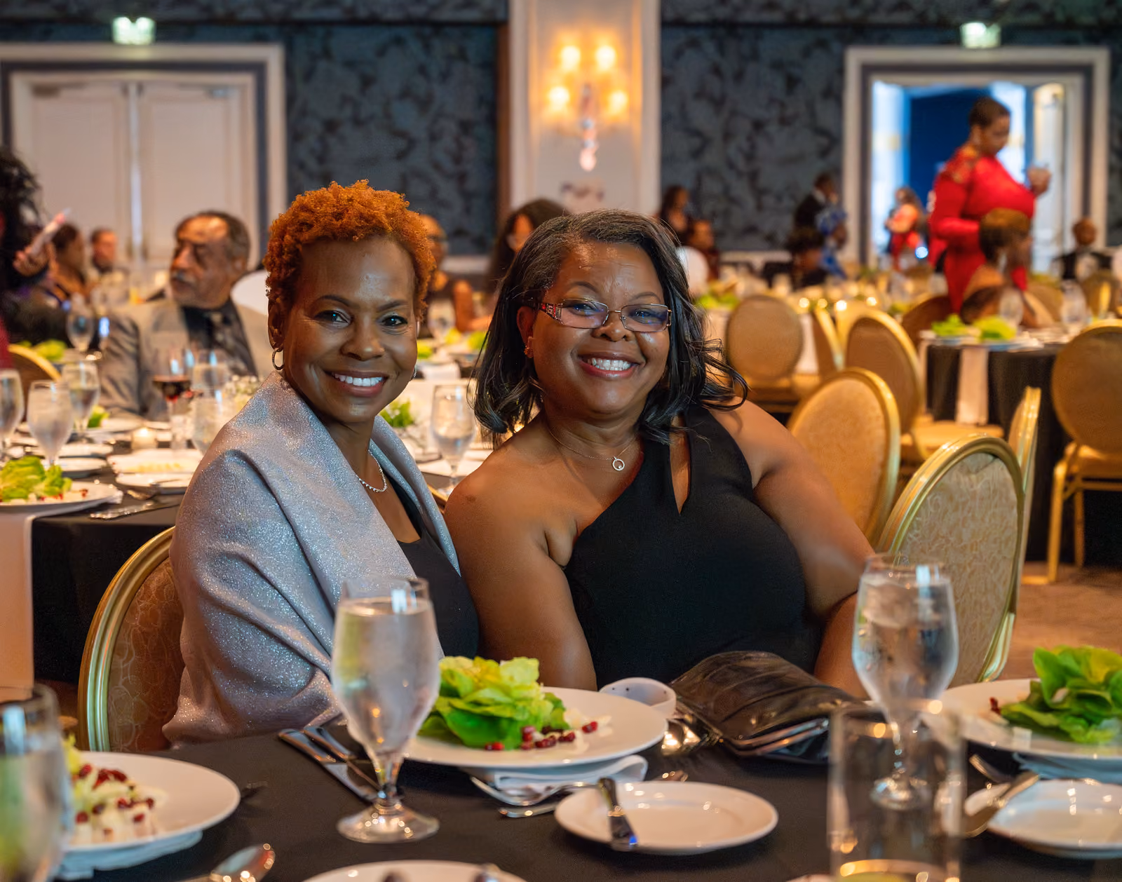 Two smiling guests sit together at a banquet table set with salads, plates and water glasses