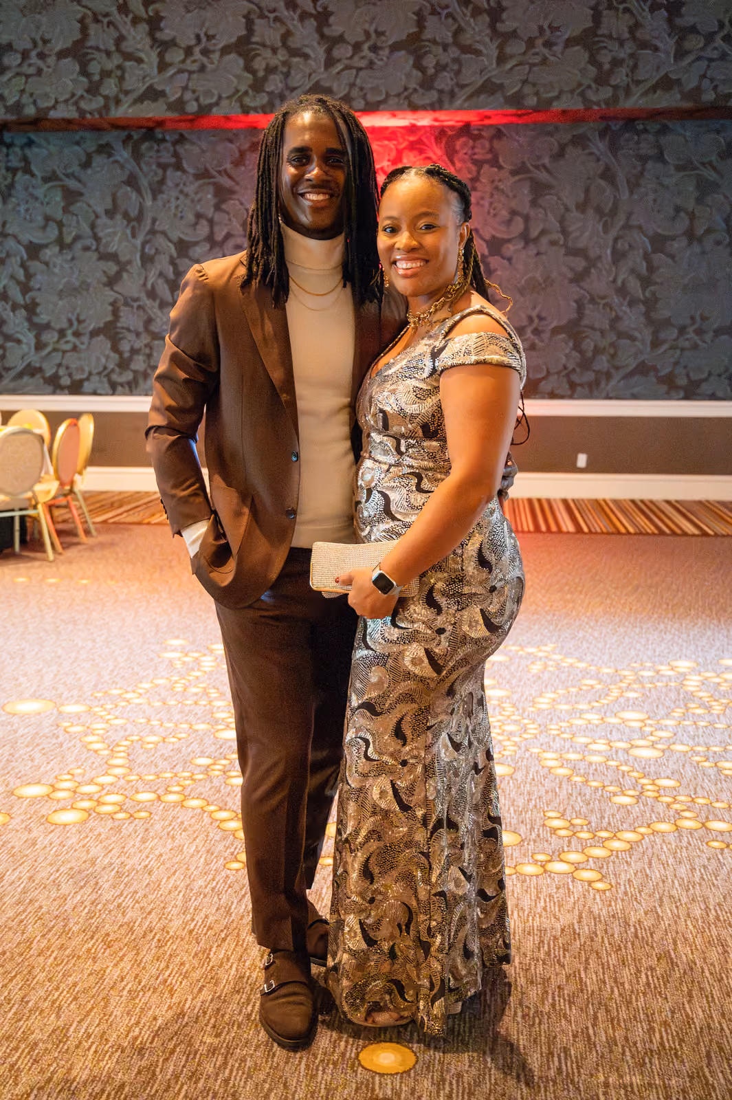 Two guests in a brown suit and a silver patterned gown pose together against a patterned ballroom wall