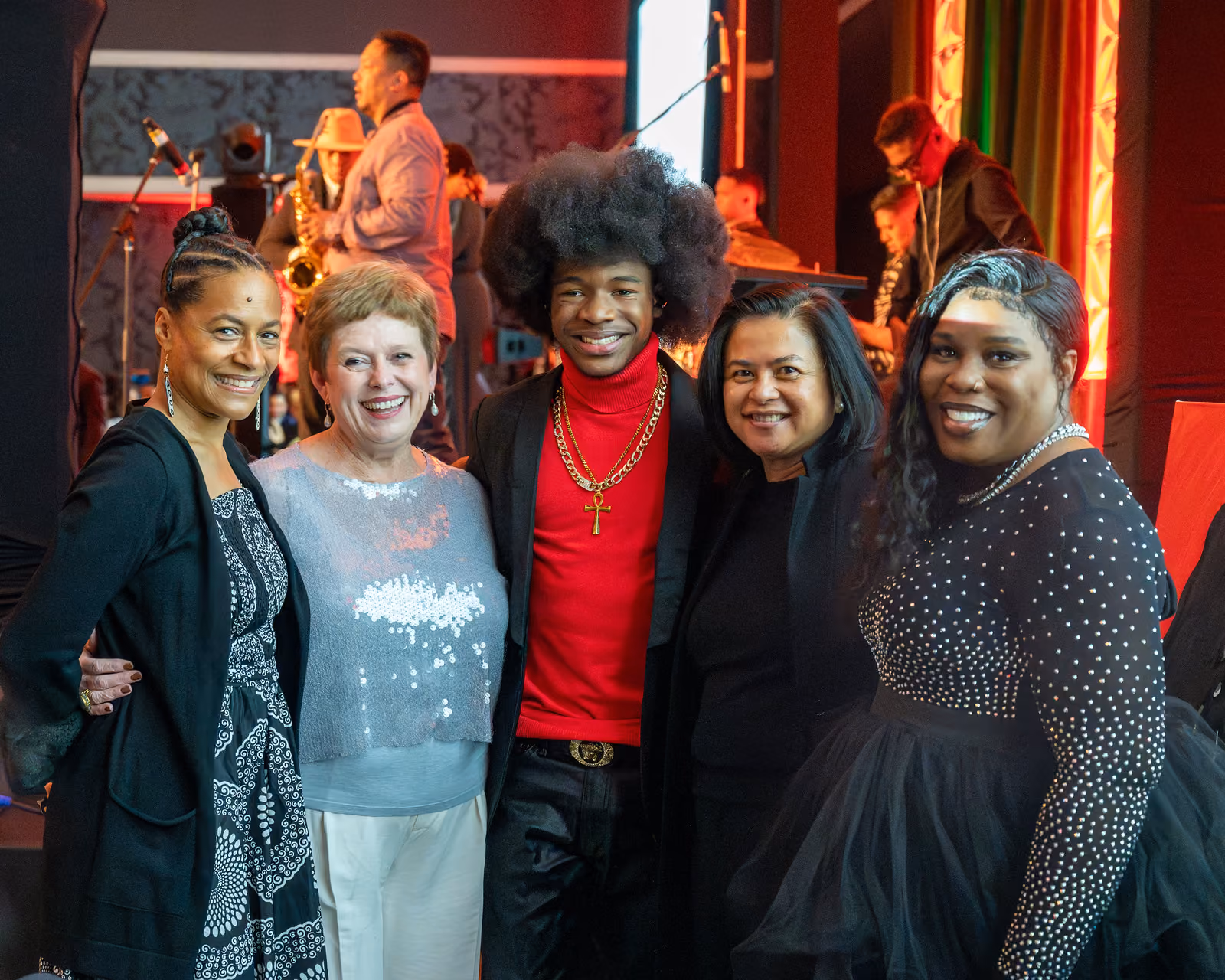 Five smiling guests pose together beside the stage while musicians perform behind them