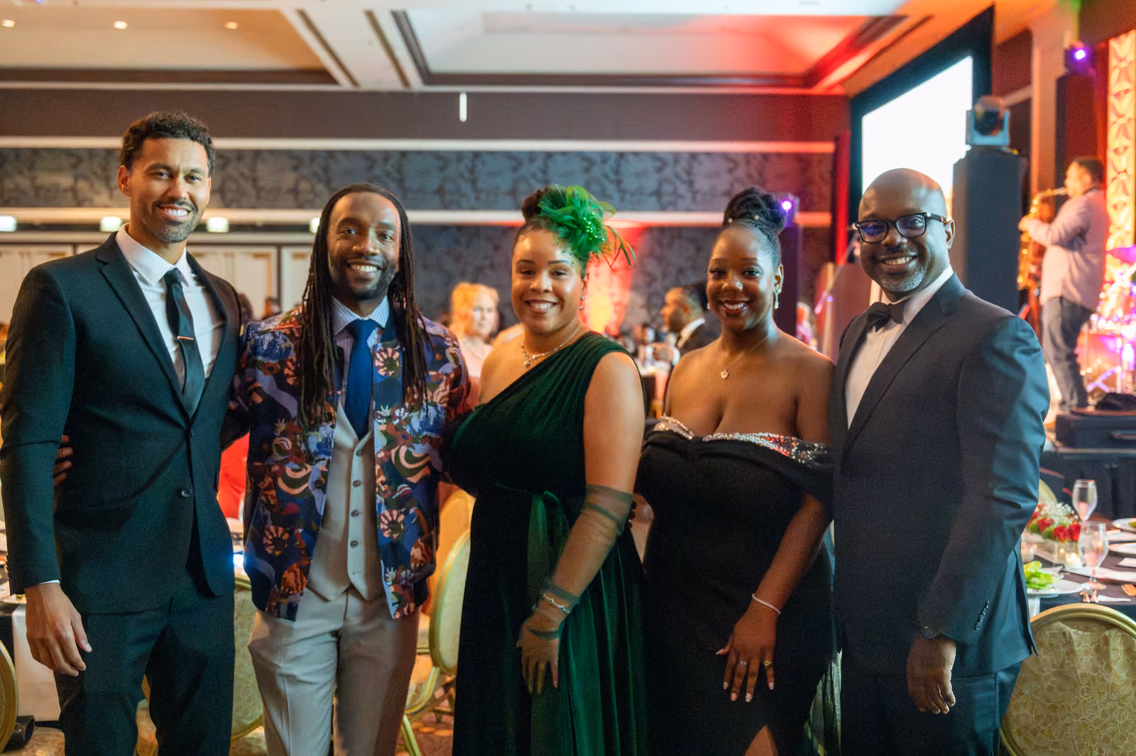 Five smiling guests in suits and evening gowns pose together near the ballroom stage