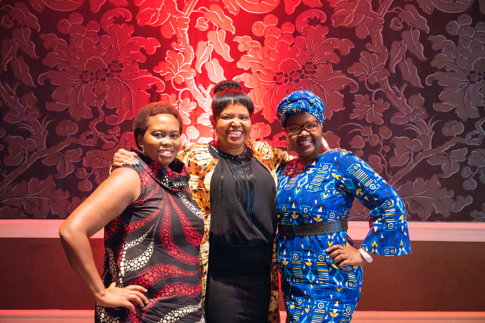 Three smiling guests in colorful patterned outfits pose with their arms around each other by a red-lit wall