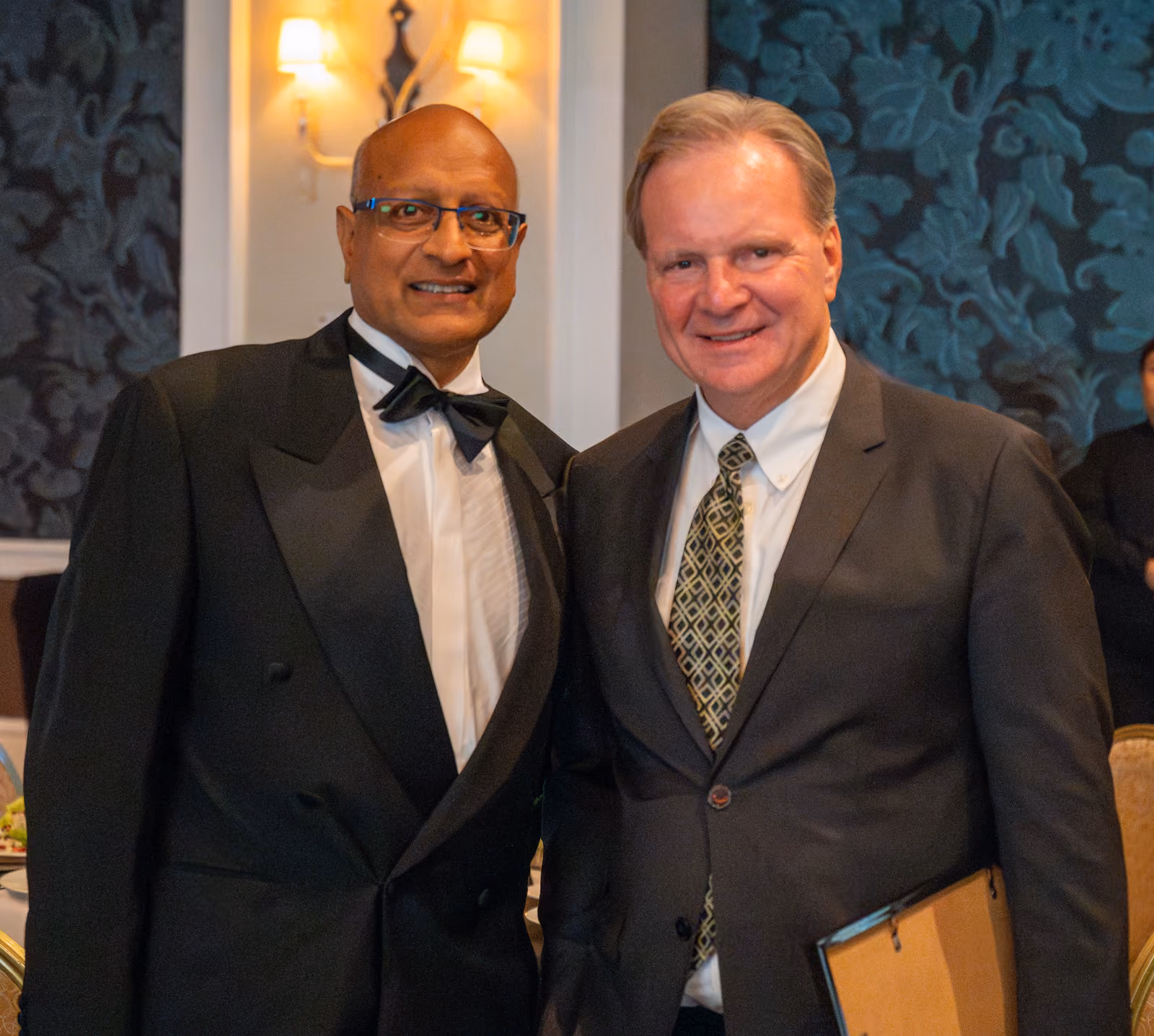 Two smiling guests in dark suits pose together in the ballroom, one holding a framed item