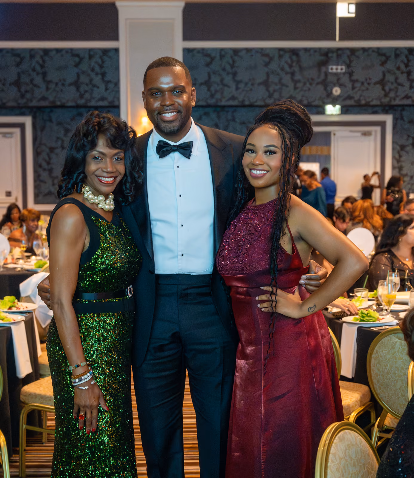 Three smiling guests in a tuxedo, green sequined gown and burgundy gown pose among banquet tables