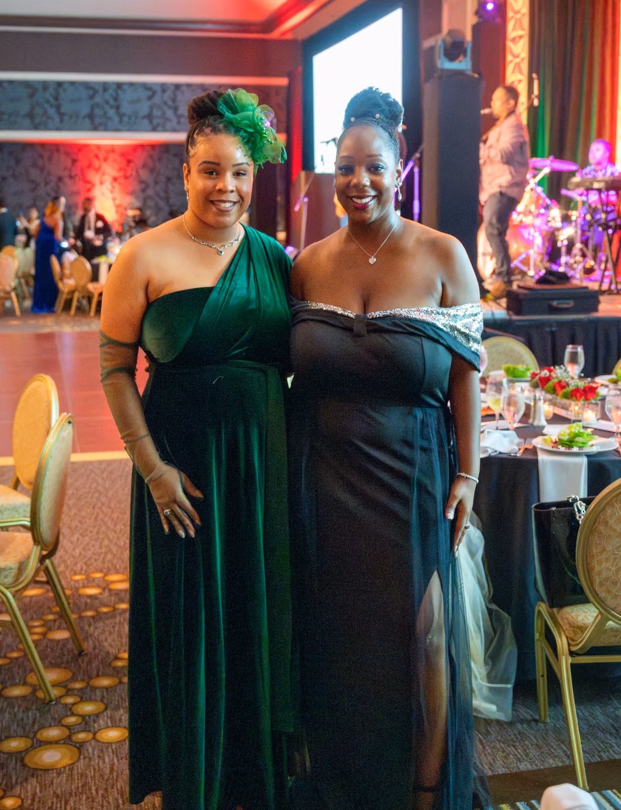 Two guests in green and black gowns pose beside banquet tables with musicians onstage behind them