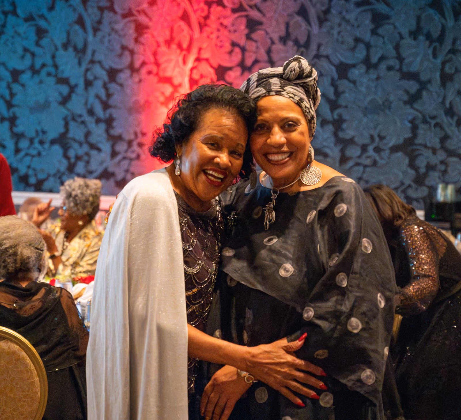 Two smiling guests, one in a silver shawl and one in a patterned headwrap, embrace beside banquet tables