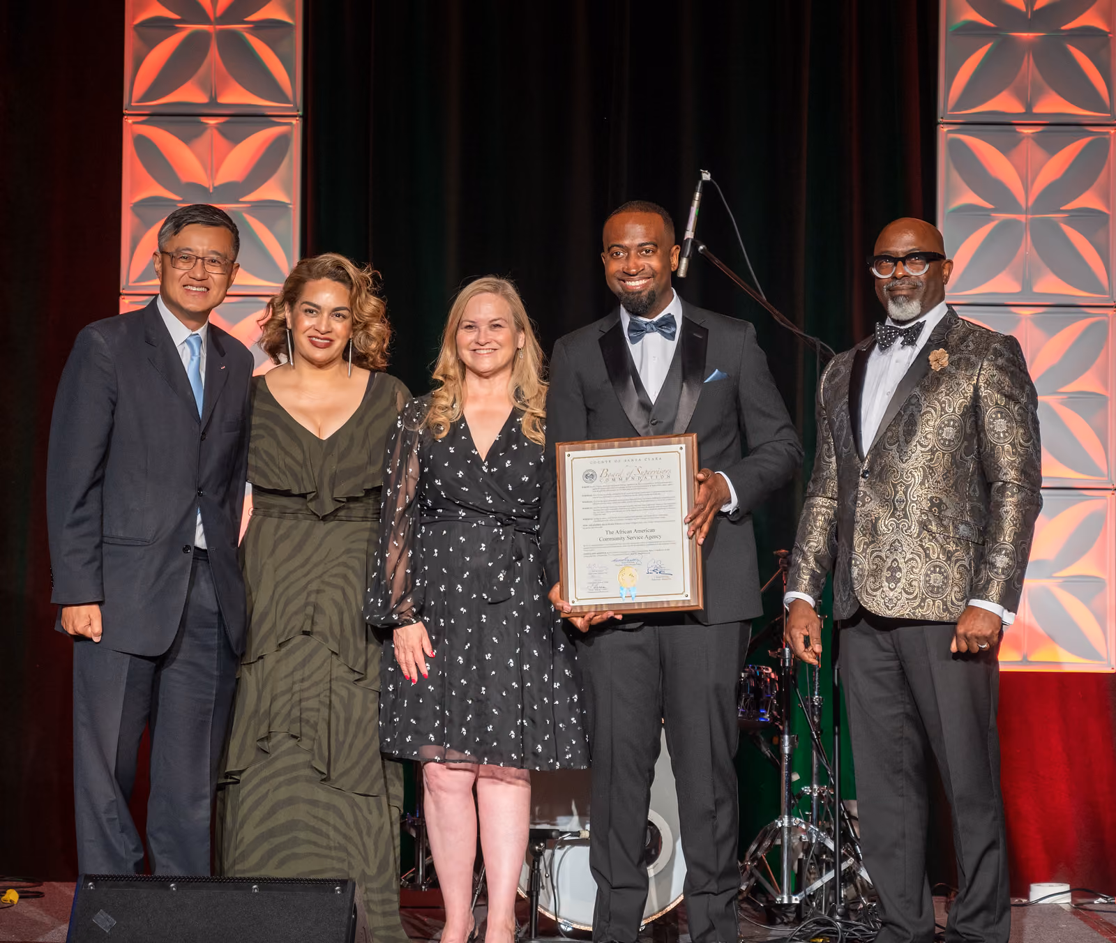 Five formally dressed guests pose onstage, with a framed certificate held at the center