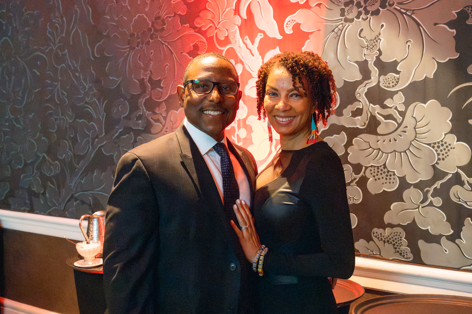 Two smiling guests in formal black attire pose together against a floral wall with red lighting