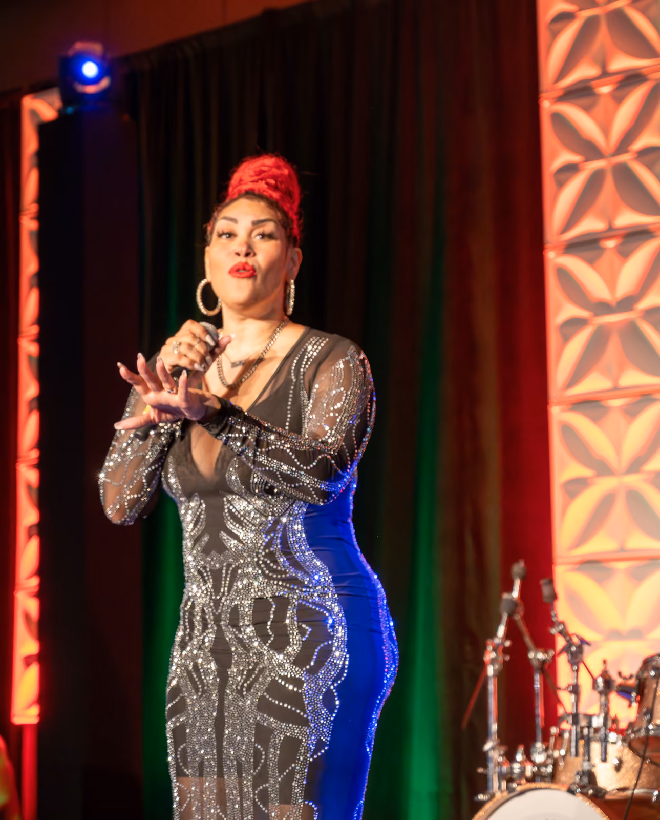 A performer with a red updo holds a microphone onstage in a sparkling black and silver gown