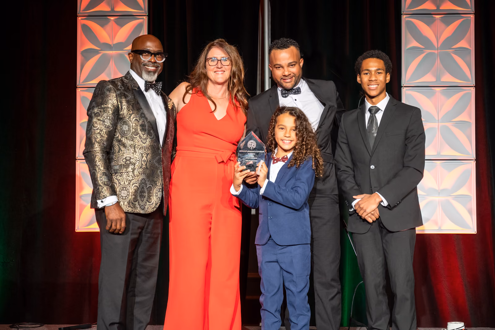 Five guests pose onstage, with a guest in a blue suit holding a clear award at the center